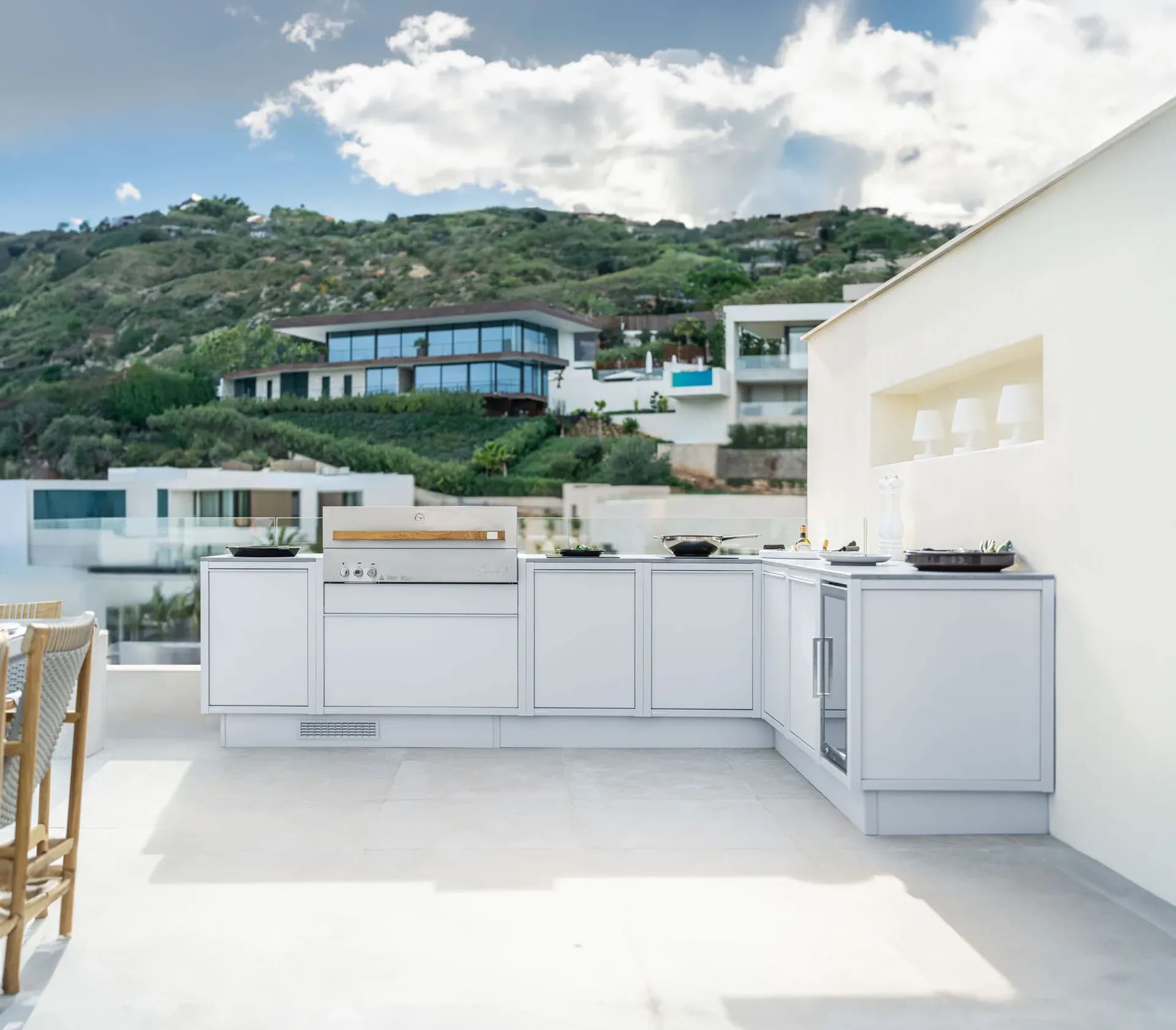 Cuisine d'extérieur claire avec grill en inox sur une terrasse ensoleillée, derrière un coteau avec des villas et la mer