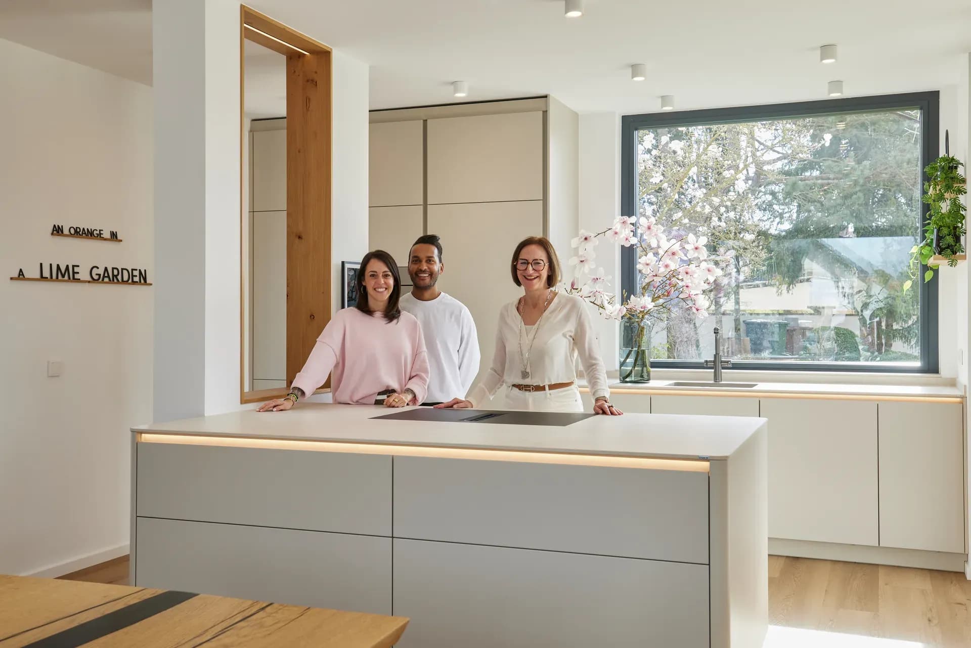 Trois personnes souriantes derrière l'îlot de cuisson gris clair d'une cuisine neuve, derrière la fenêtre un magnolia en fleurs