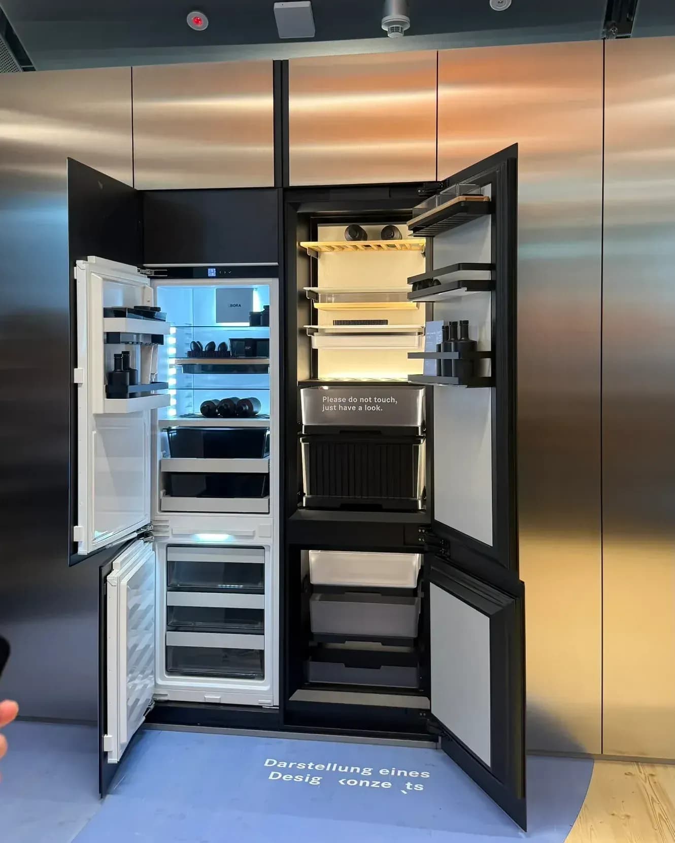 Au BORA Flagship Store de Herford, à la Küchenmeile 2026 : réfrigérateurs et congélateurs ouverts, intégrés dans un mur en inox et signalés au sol comme présentation d’un concept de design.