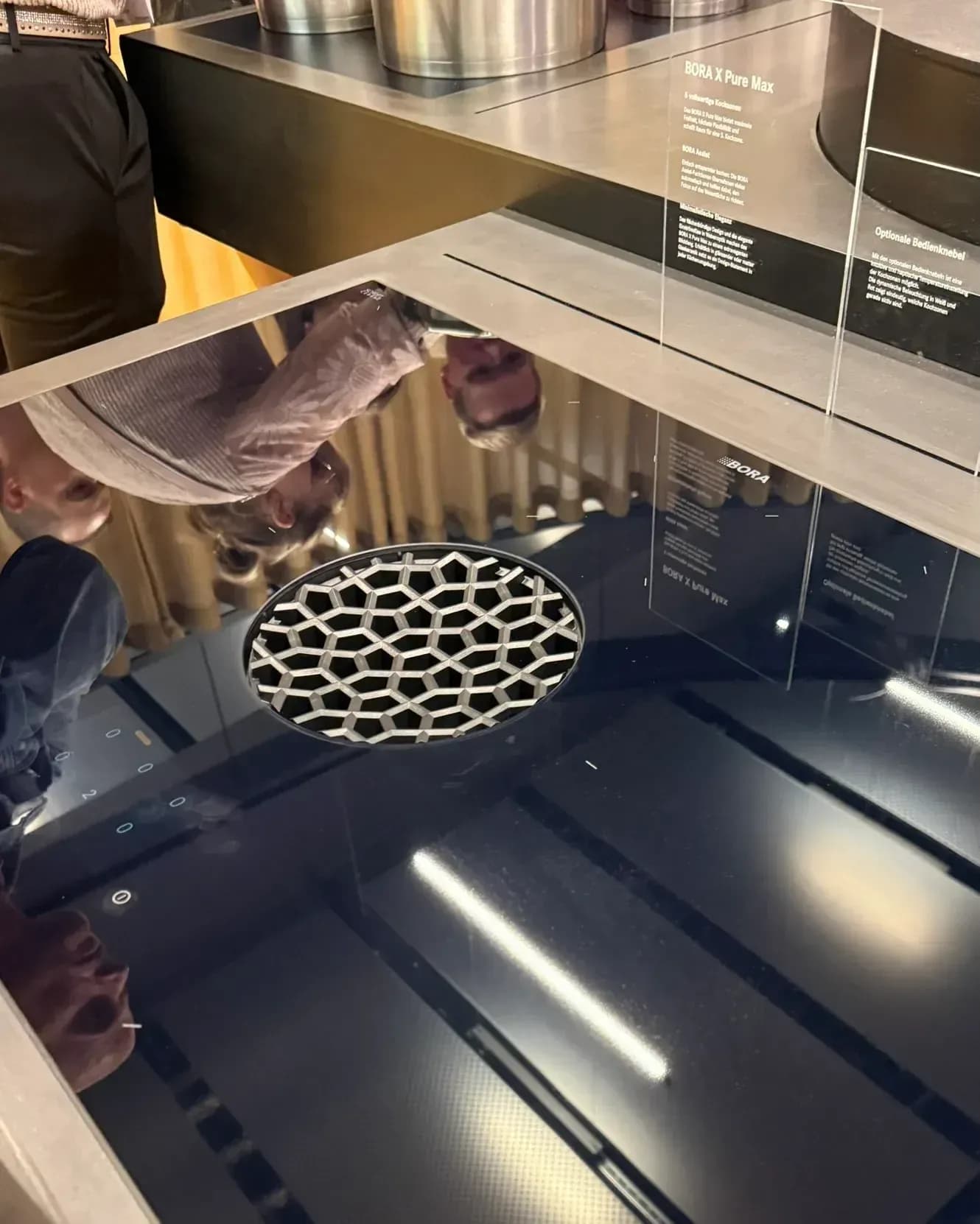 Au BORA Flagship Store de Herford, à la Küchenmeile 2026 : table de cuisson aspirante BORA X Pure Max avec ouverture d’aspiration ronde dans la surface vitrocéramique noire, derrière un panneau d’information.