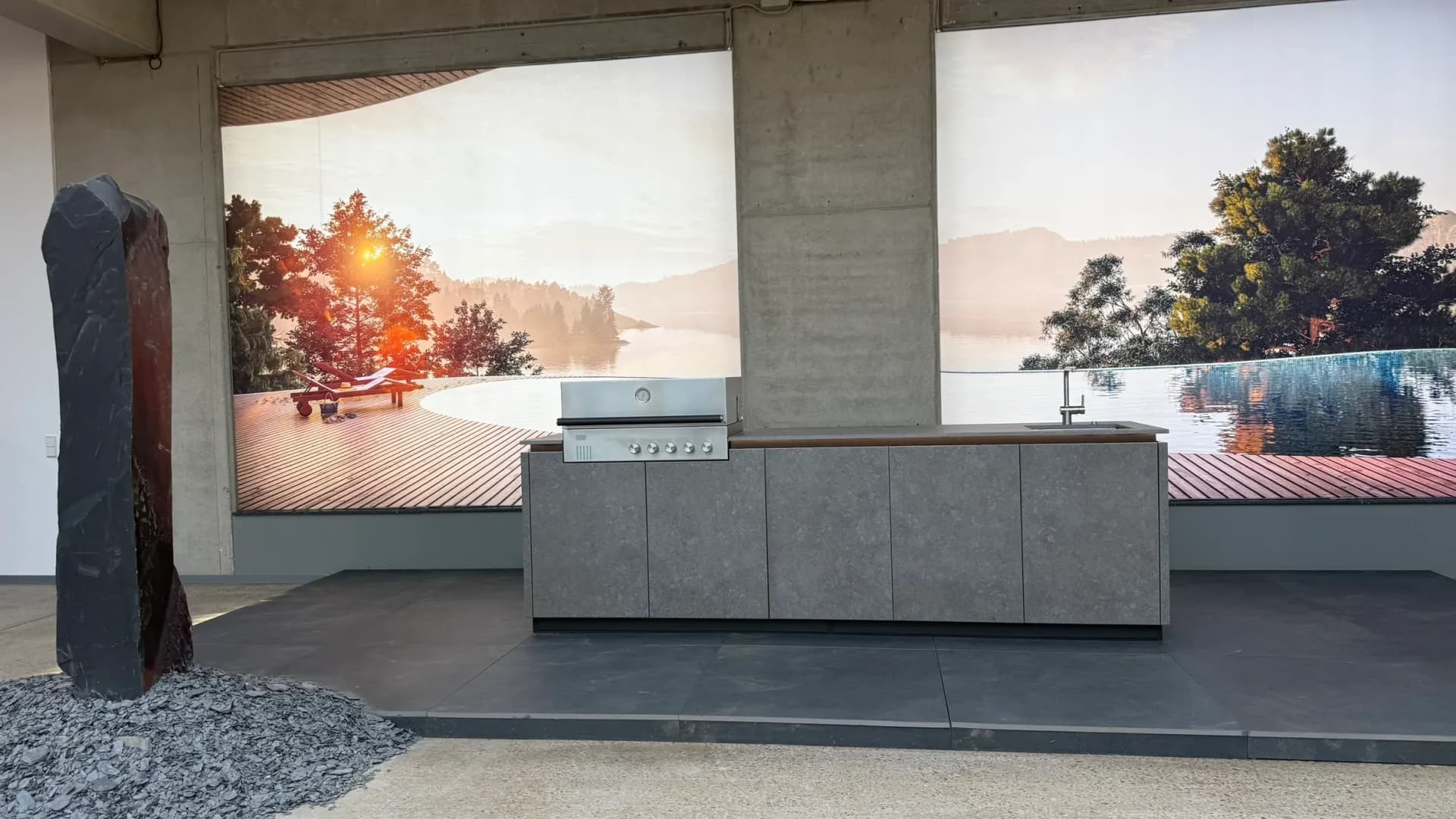 Chez BURN Outdoor Kitchen à Bissendorf, à la Küchenmeile 2026 : îlot de cuisine extérieure à façades grises avec barbecue en inox et évier, devant un grand papier peint photo de paysage.