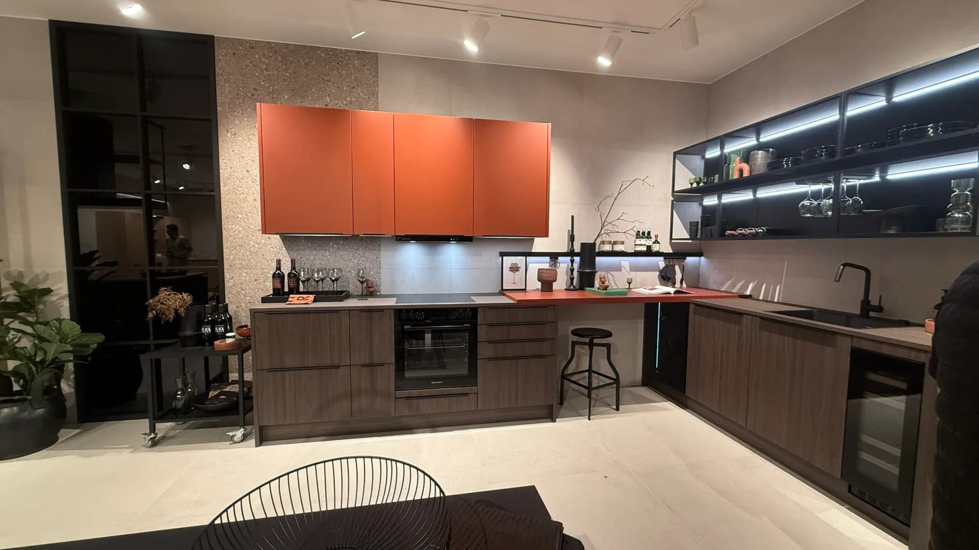 Dans l’exposition de Häcker Küchen, à la Küchenmeile 2026 : cuisine en L avec éléments hauts couleur terre cuite au-dessus d’éléments bas en décor bois foncé, à droite une étagère murale ouverte éclairée.