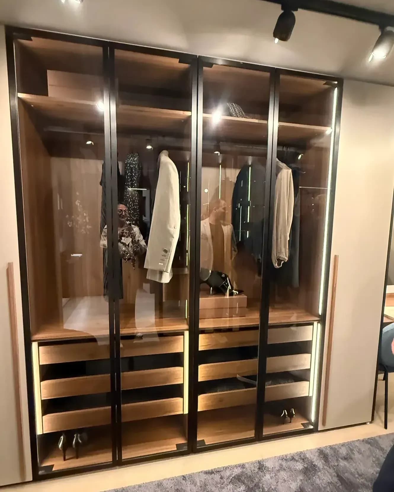 Dans l’Architekturwerkstatt de LEICHT à Löhne, à la Küchenmeile 2026 : armoire-dressing à portes vitrées cadrées de noir, aménagement intérieur en bois, tiroirs éclairés et penderies.