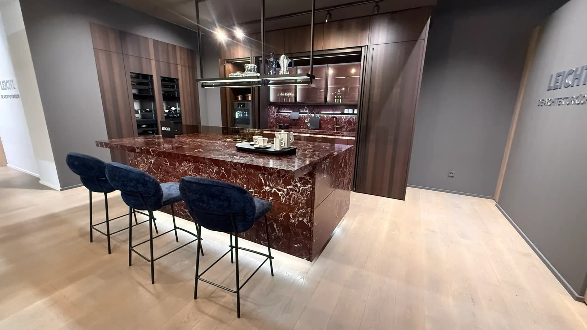 Dans l’Architekturwerkstatt de LEICHT à Löhne, à la Küchenmeile 2026 : îlot habillé de marbre rouge, plan de travail compris, devant lui trois tabourets de bar tapissés de bleu, derrière des armoires toute hauteur en bois foncé et une niche avec vitrines.