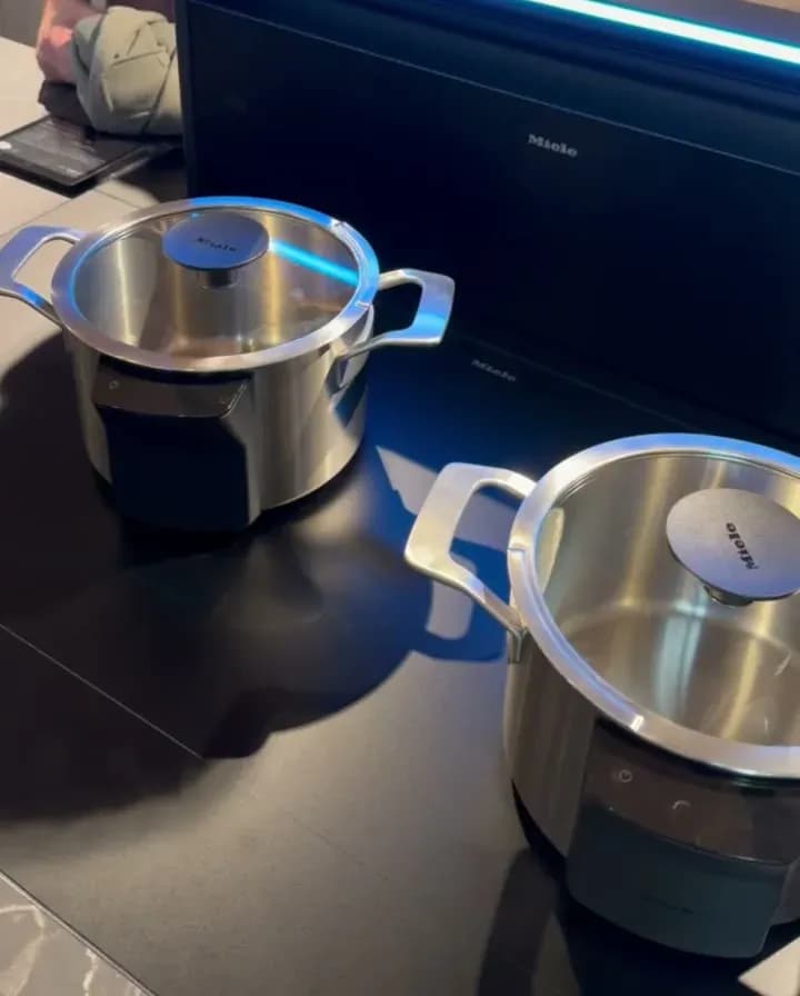 Stand Miele à la Küchenmeile 2026 : deux casseroles en inox sur une table de cuisson noire, derrière une hotte escamotable relevée au bord éclairé en bleu.