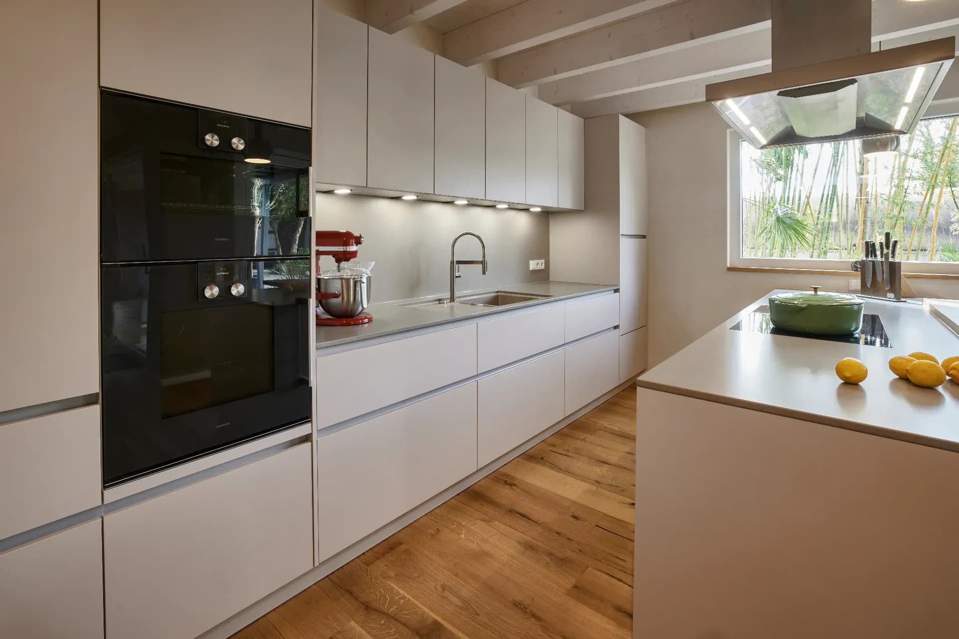 Cuisine linéaire à façades greige sans poignées, fours Gaggenau et îlot de cuisson sous une hotte en inox
