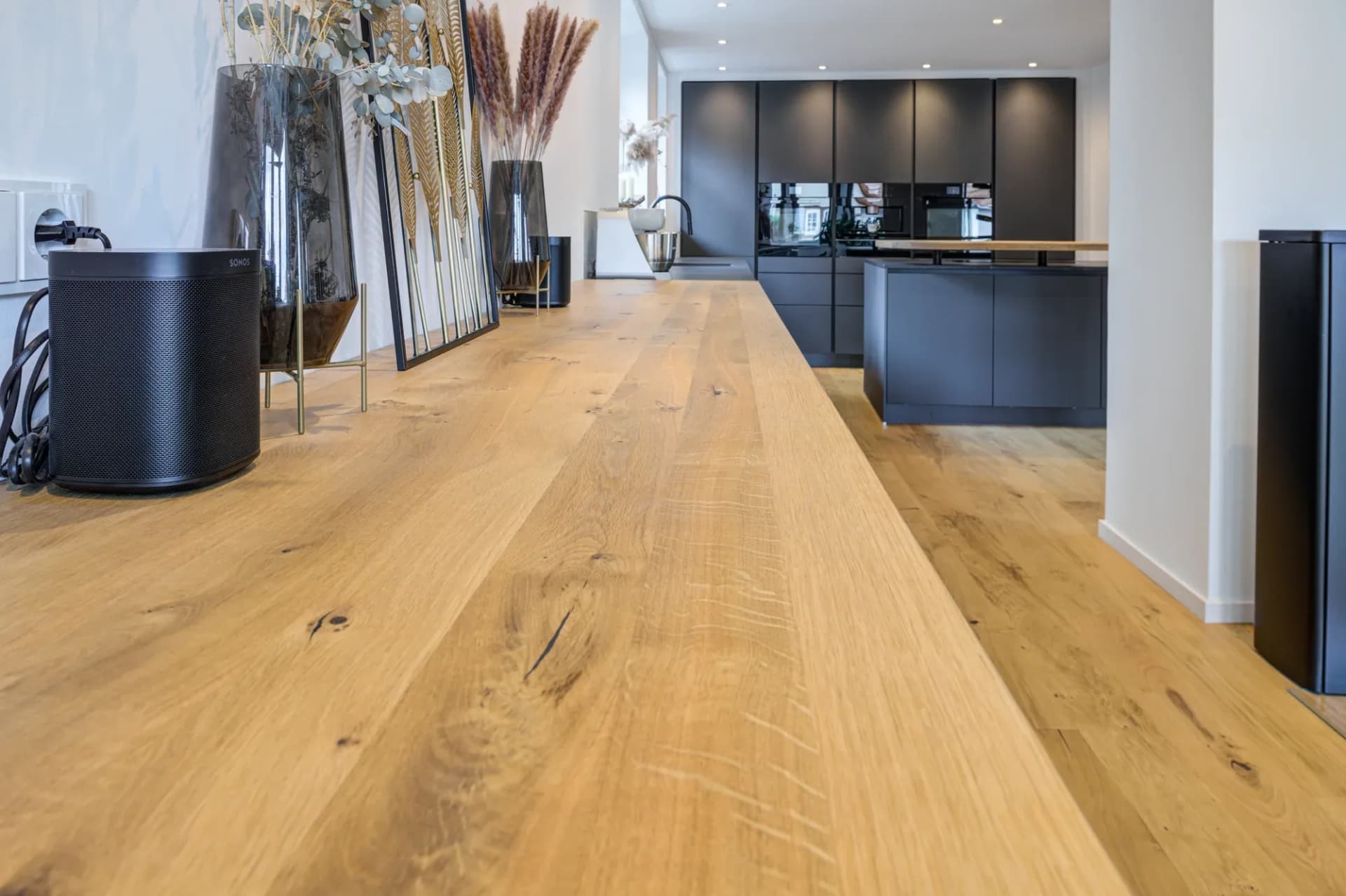 Un parquet en chêne rustique huilé s'étend en perspective vers la cuisine mate et sombre