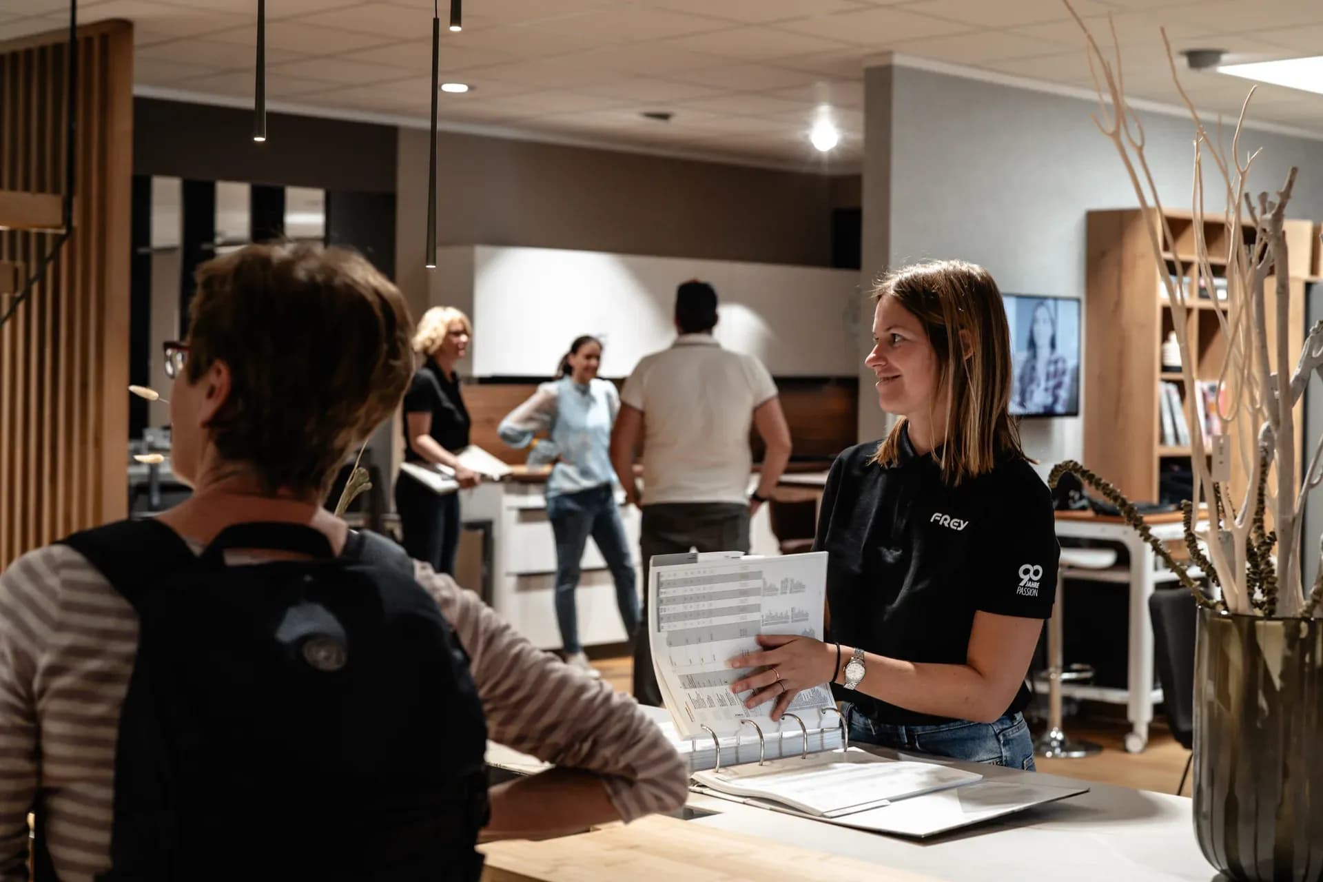 Conseillère Frey montrant des documents dans un classeur à une visiteuse, derrière des visiteurs entre les cuisines du showroom de Kandel