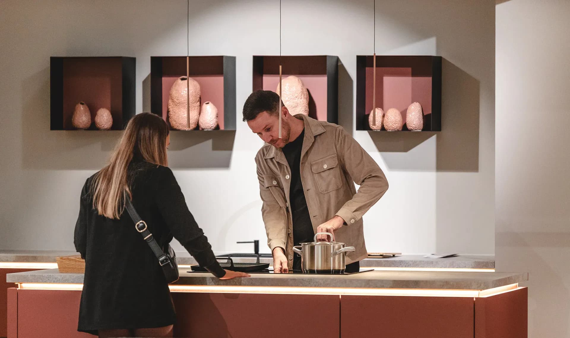 Deux visiteurs à l'îlot de cuisson d'une cuisine d'exposition rouge, une casserole sur la plaque, derrière des caissons muraux avec des vases