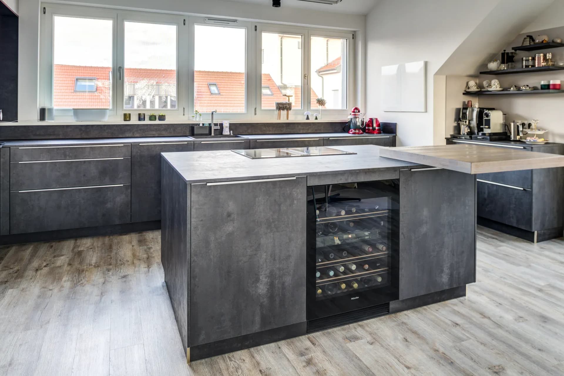 Cuisine ouverte sous les combles avec finition béton : îlot de cuisson gris indépendant avec plan de bar en chêne rabattable, colonnes blanches avec niche ouverte en arrière-plan, sol en bois.