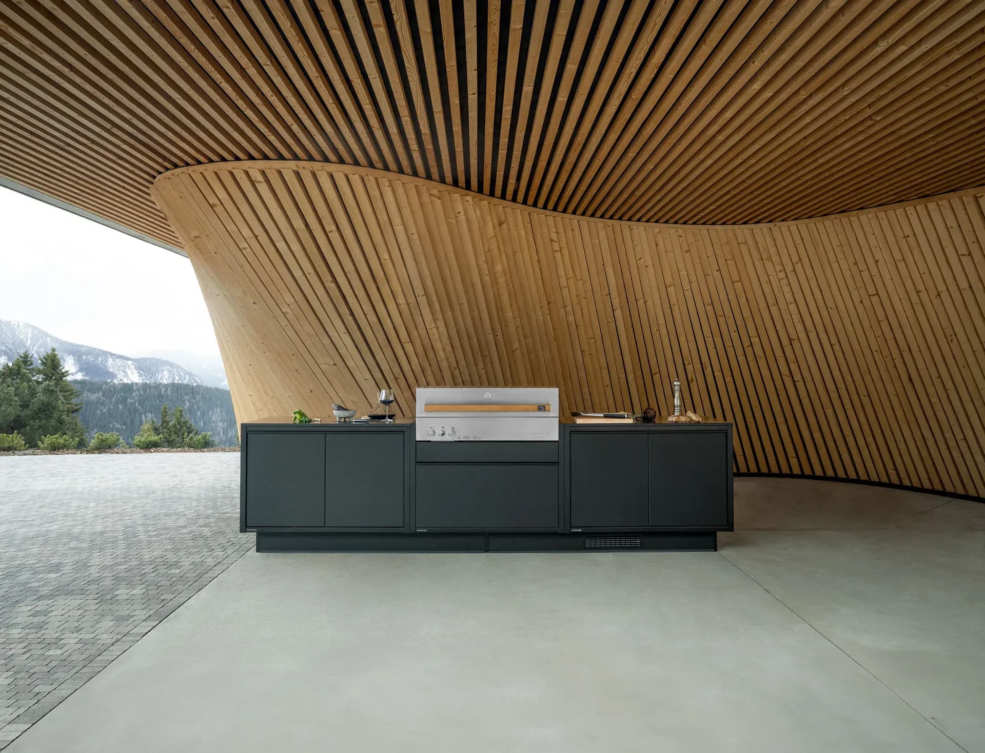 Cuisine d'extérieur BURN anthracite avec grill en inox sous un auvent galbé à lames de bois, montagnes enneigées à l'arrière-plan