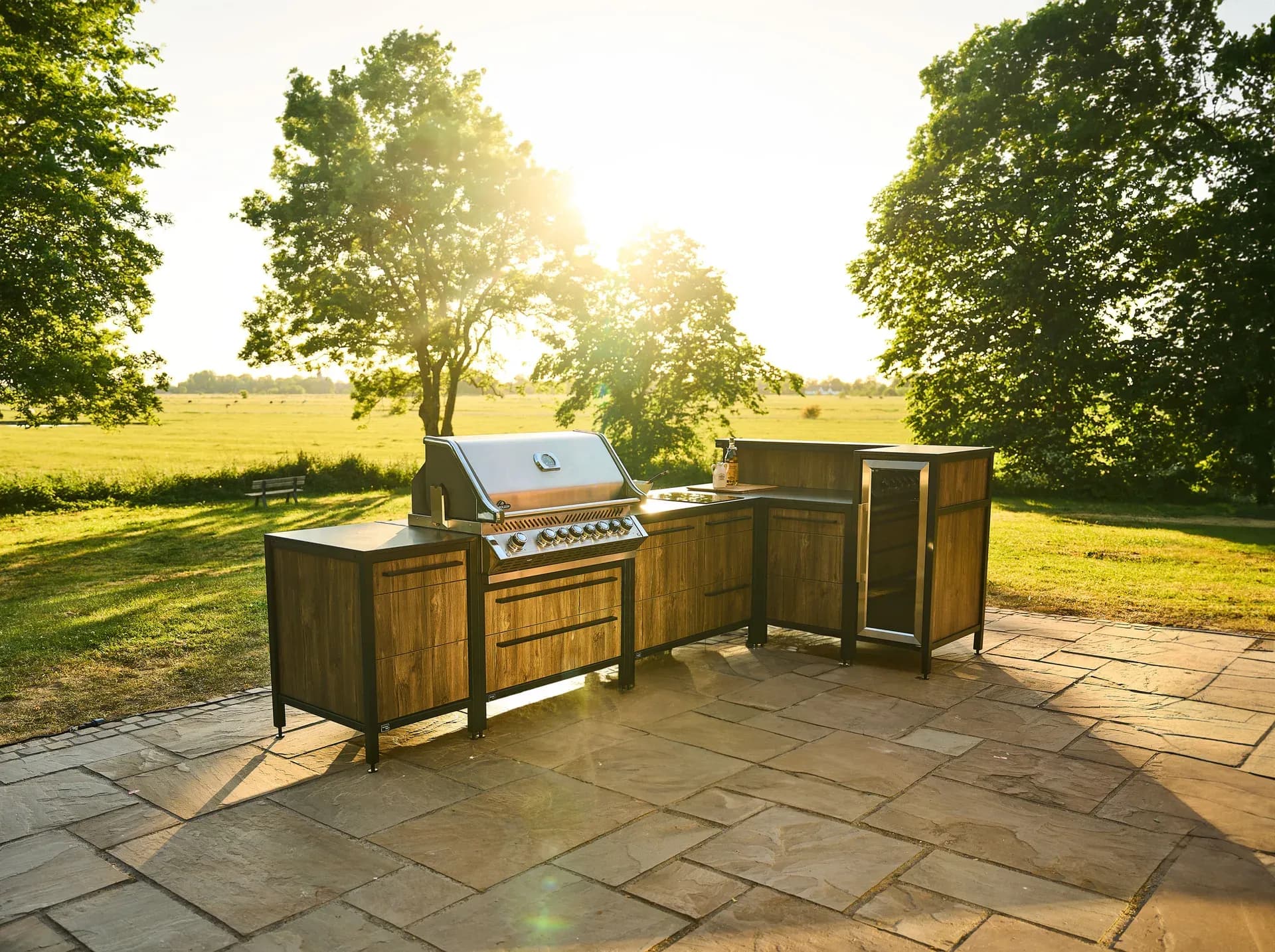 Cuisine d'extérieur en angle avec façades en bois et grill en inox sur une terrasse en pierre naturelle, derrière une prairie et des arbres dans la lumière du soir