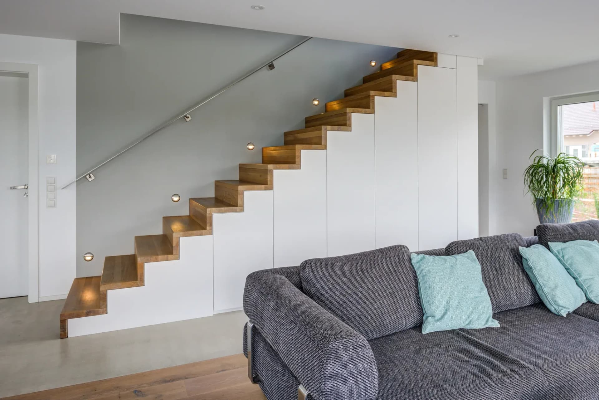 Séjour ouvert avec escalier plié en chêne qui s'élève au-dessus d'un mur d'armoires blanc fermé, appliques encastrées et canapé gris.