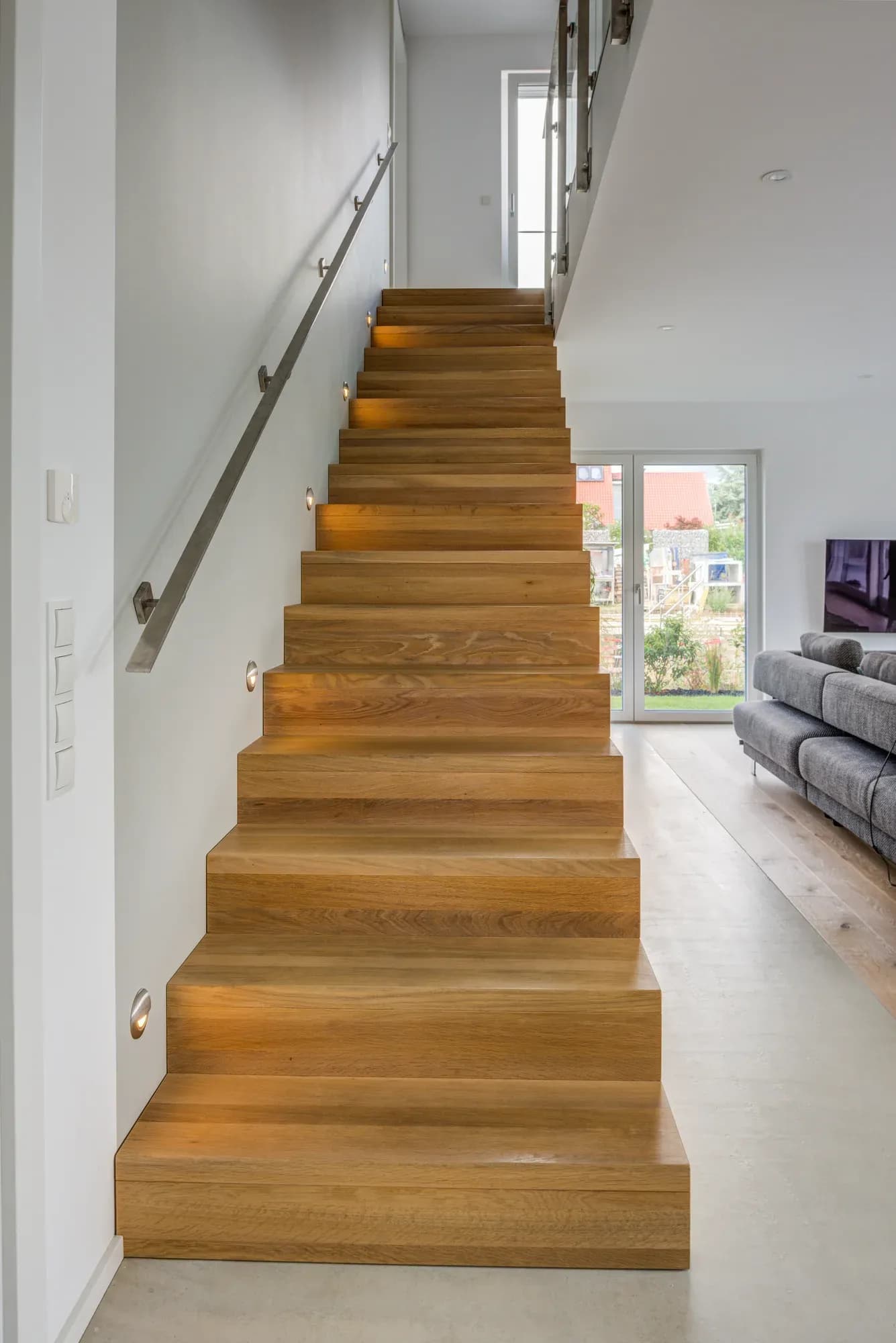Escalier plié en chêne massif qui monte vers la lumière du jour, avec main courante inox au mur et vue sur le séjour attenant.