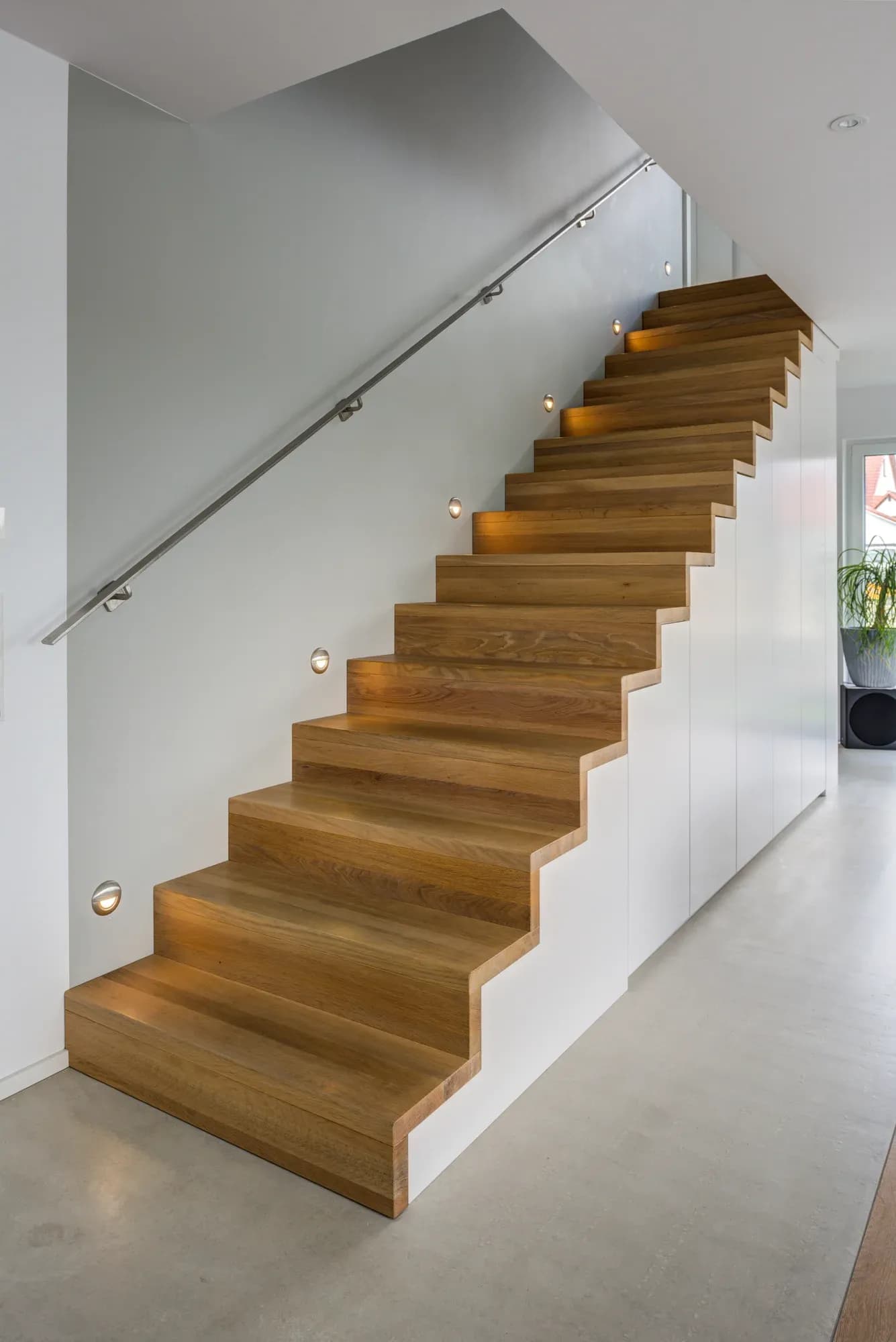 Escalier plié en chêne au-dessus de façades d'armoires blanches sans poignées, accompagné d'appliques rondes éclairant les marches.
