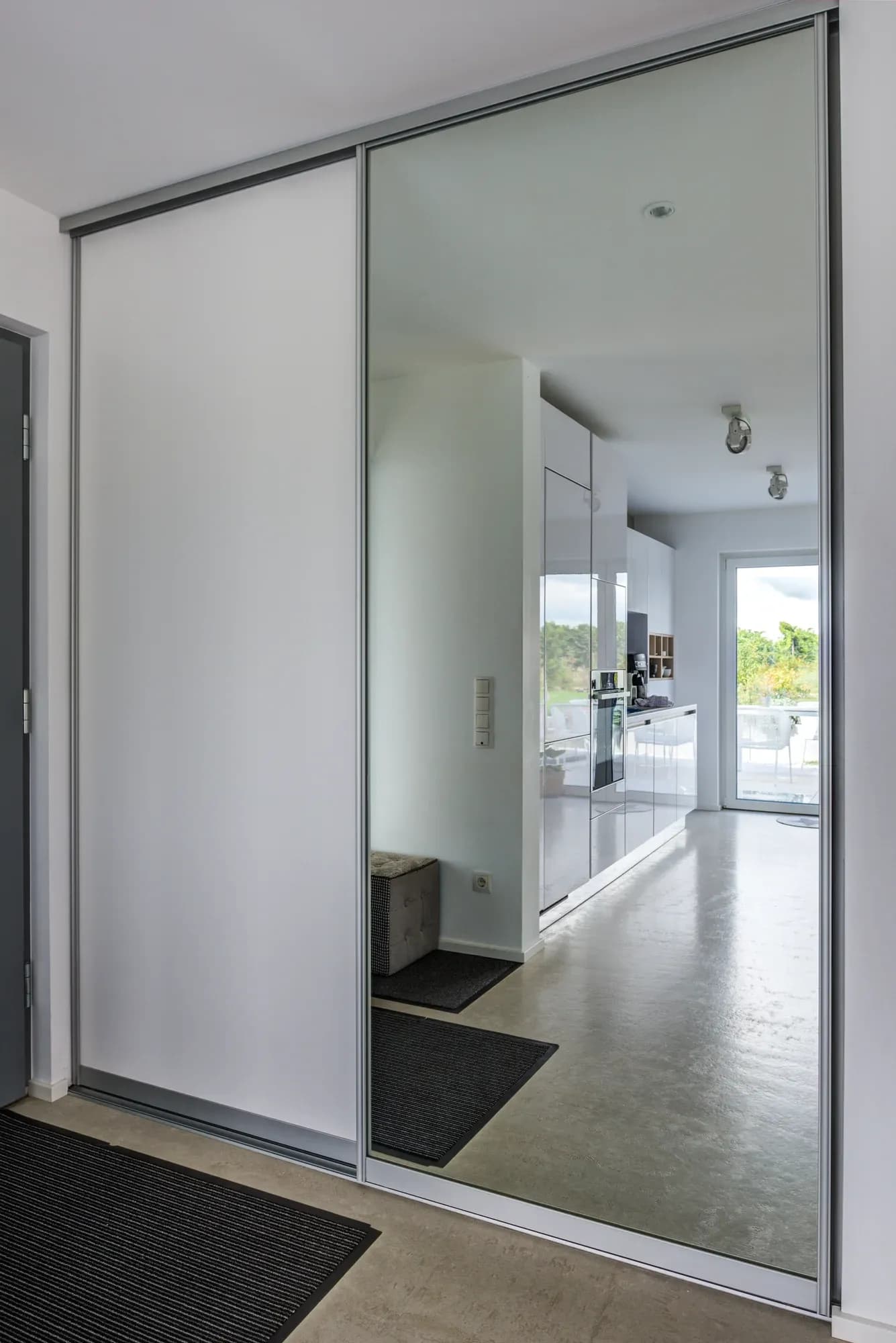Porte coulissante miroir de l'armoire d'entrée, où se reflètent la cuisine blanche ouverte avec four encastré et la sortie vers la terrasse.