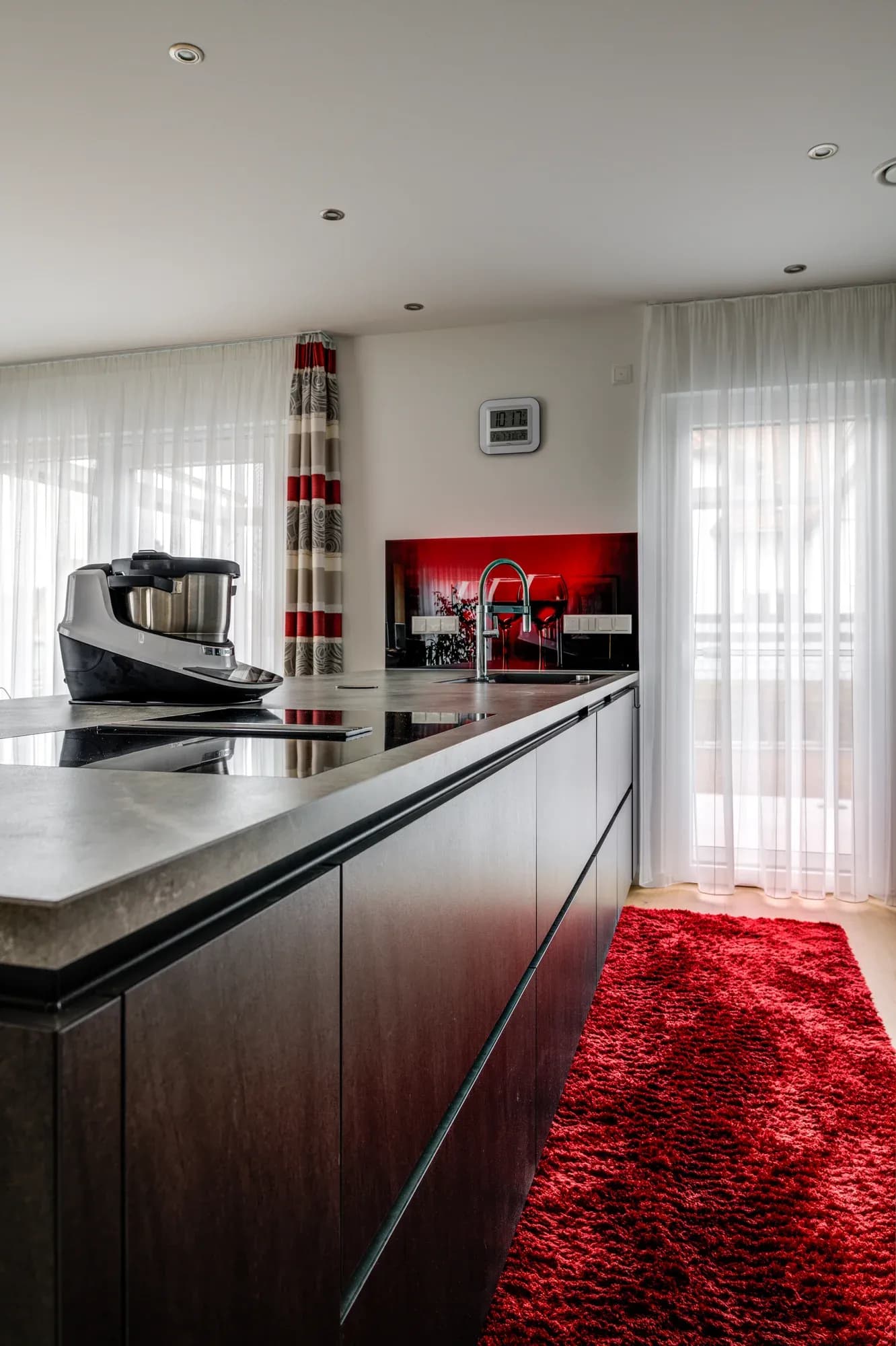 Rangée de cuisine blanche sans poignées avec îlot de cuisson foncé, tapis de couloir rouge et crédence en verre rouge, lumière du jour provenant de deux fenêtres
