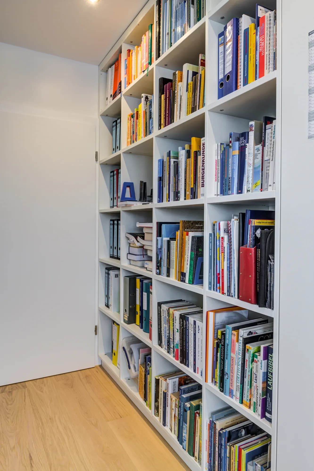 Bibliothèque murale blanche toute hauteur sur parquet en chêne, remplie de livres triés par couleur