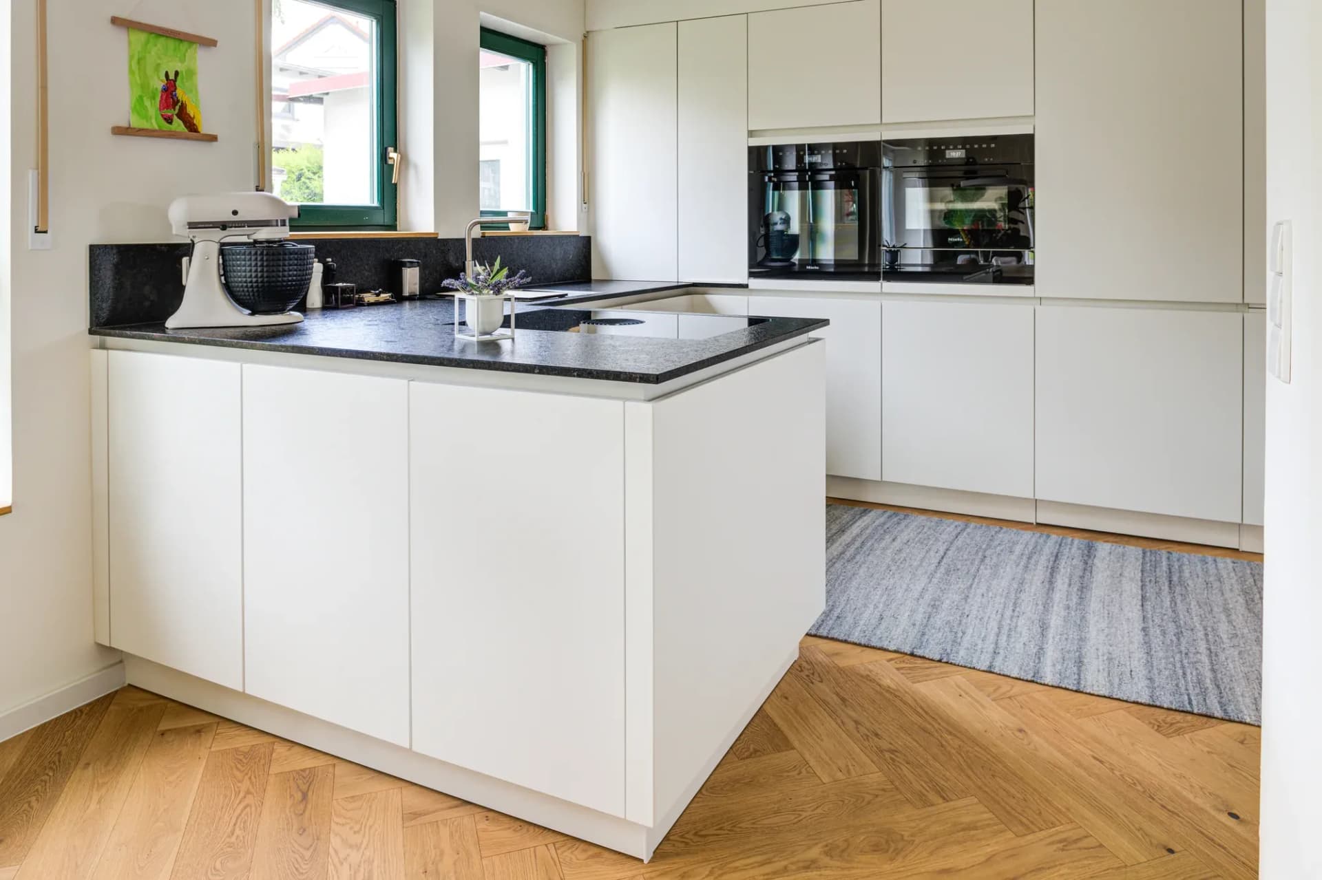 Vue depuis la porte sur la cuisine blanche en U, avec finition en pierre naturelle sombre, fenêtres en bois vertes et parquet chêne à bâtons rompus.