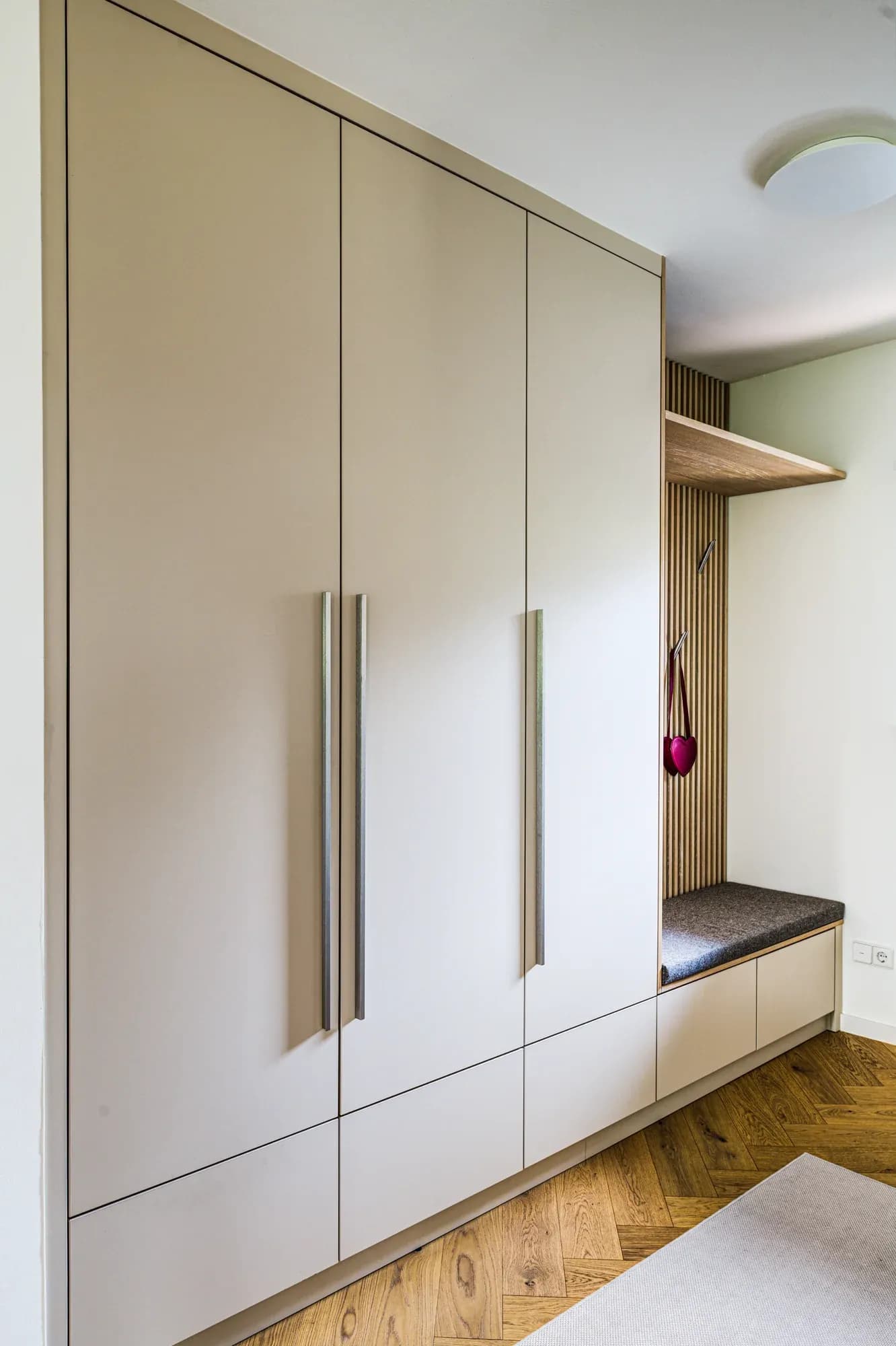 Mur de vestiaire beige chaud avec niche ouverte en lamelles de chêne, banc à assise en feutre et patères murales.