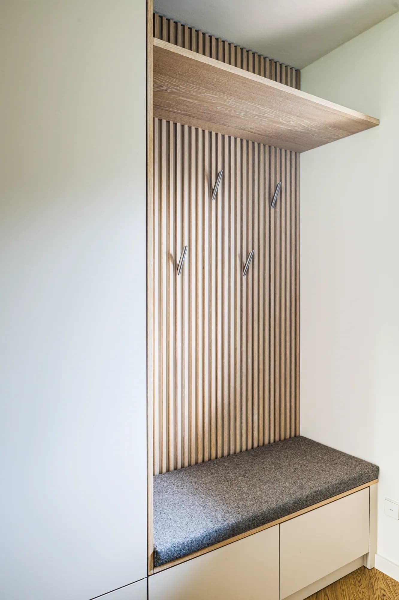 Détail de la niche du vestiaire : lamelles verticales en chêne avec patères métalliques au-dessus d'un banc à assise en feutre gris.