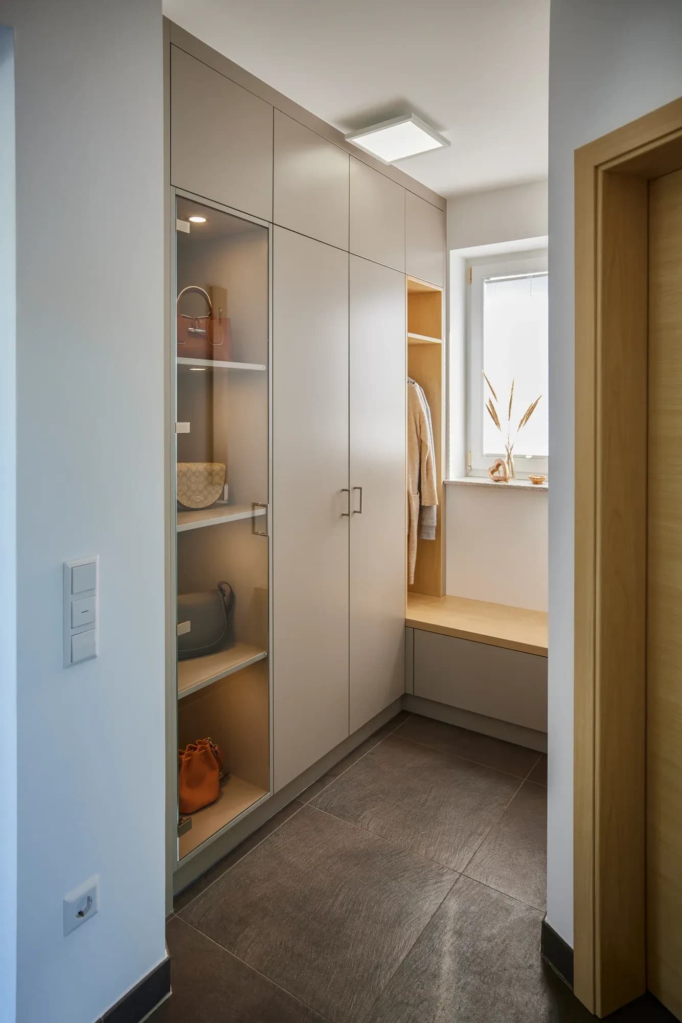 Couloir étroit avec armoire intégrée toute hauteur gris clair, vitrine ouverte éclairée, banc en bois sous la fenêtre et porte intérieure en bois clair à droite.