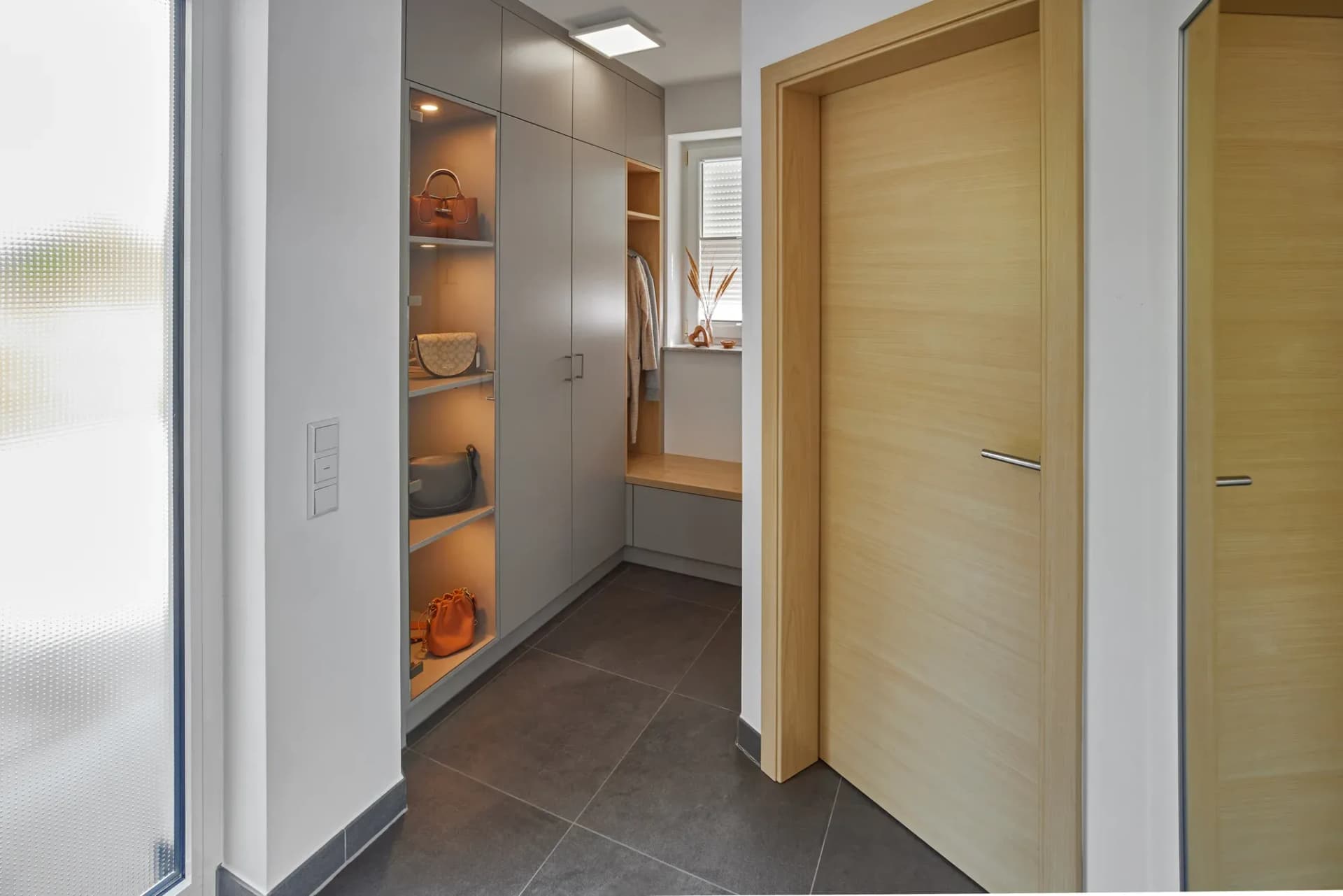Porte intérieure en bois clair à poignée fine dans le couloir ; derrière, l'armoire de vestiaire gris clair avec niche éclairée.