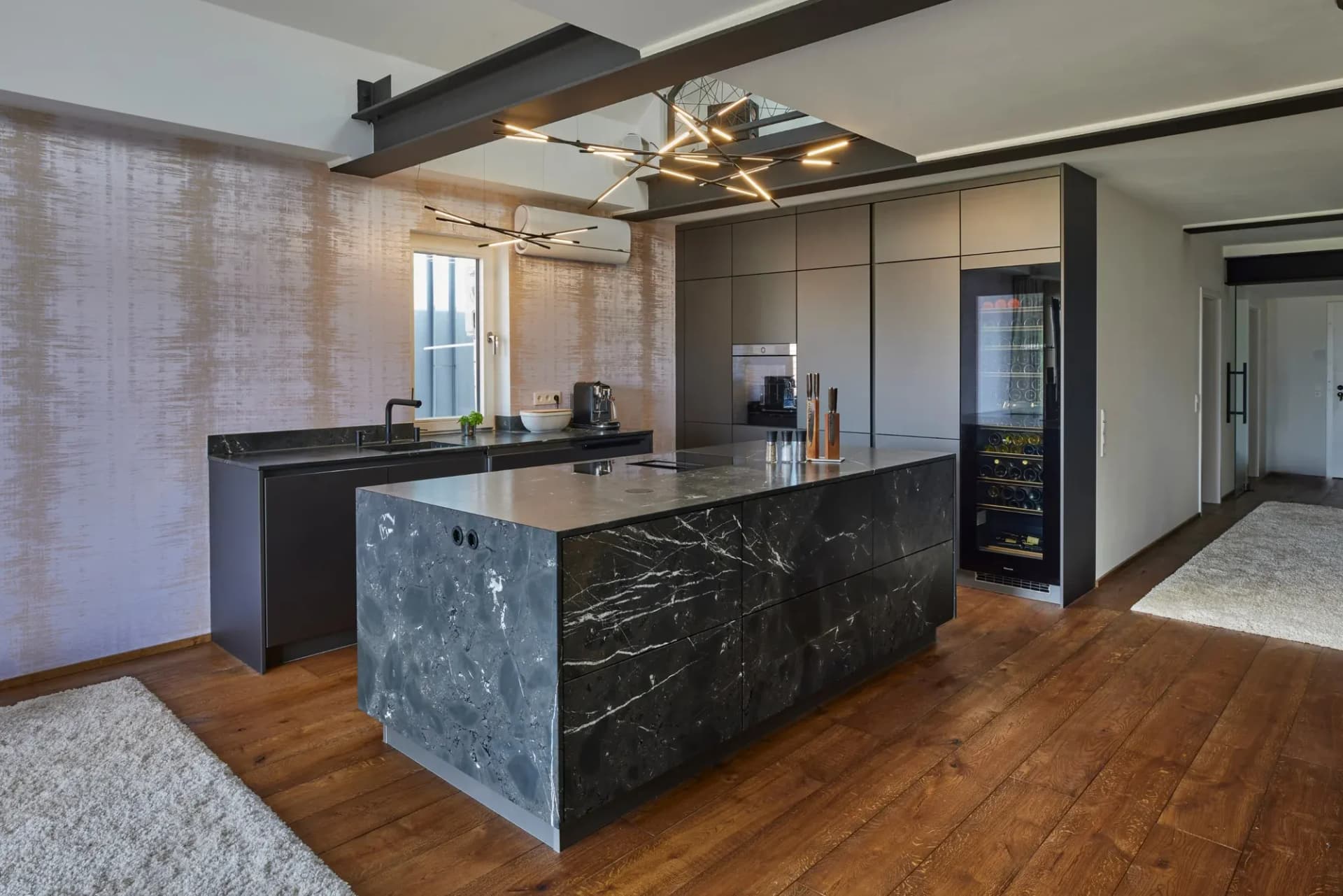 Cuisine indépendante dans un loft : îlot en pierre naturelle noire veinée de blanc, derrière un mur d'armoires gris mat avec cave à vin intégrée, sous des poutres en acier apparentes et une suspension ramifiée au-dessus d'un large plancher en chêne.