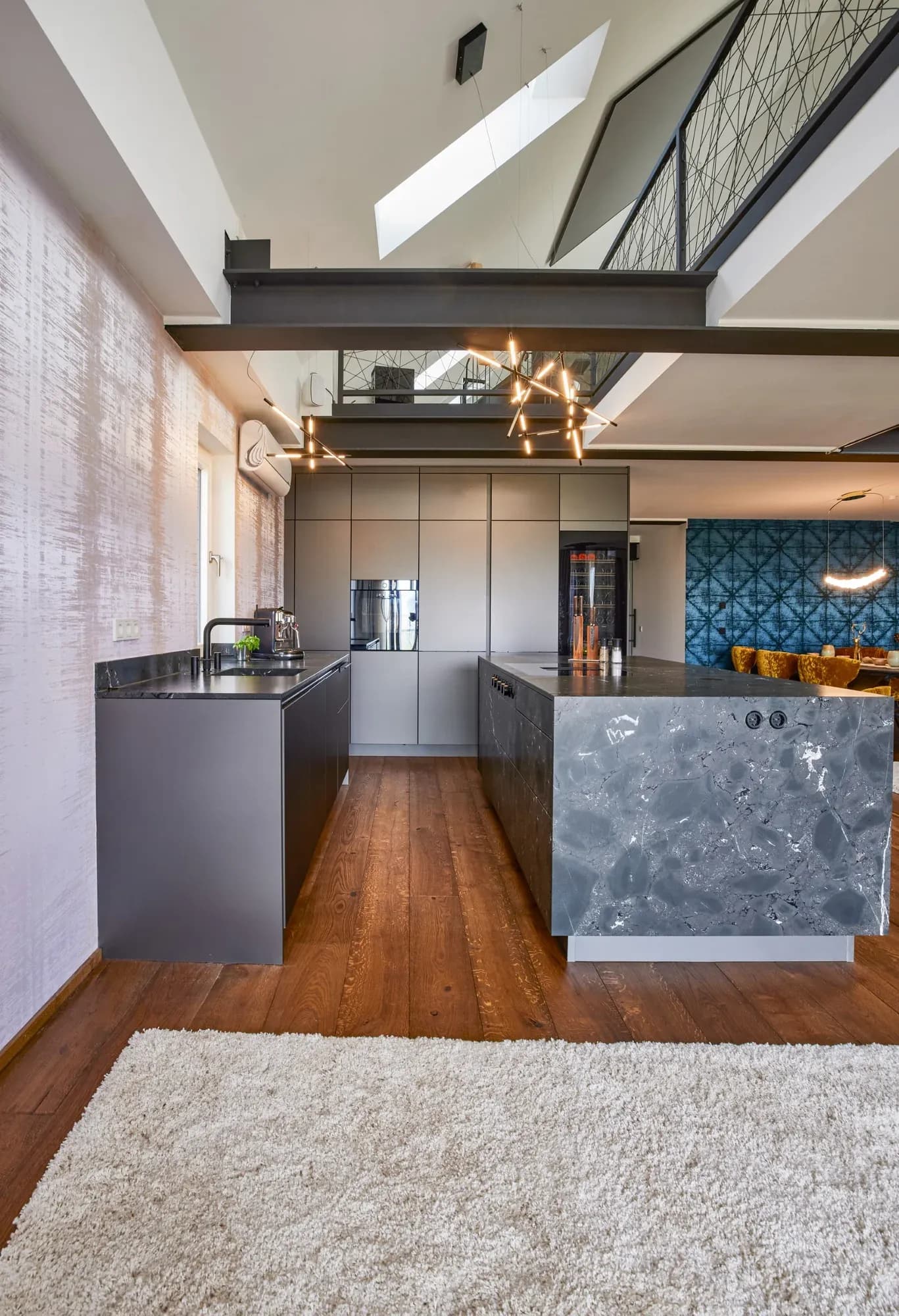 Vue à travers le loft sur deux niveaux : poutres en acier, fenêtre de toit et mezzanine au garde-corps à câbles au-dessus de la cuisine à îlot en pierre naturelle ; à droite, un coin repas aux chaises en velours jaune moutarde.