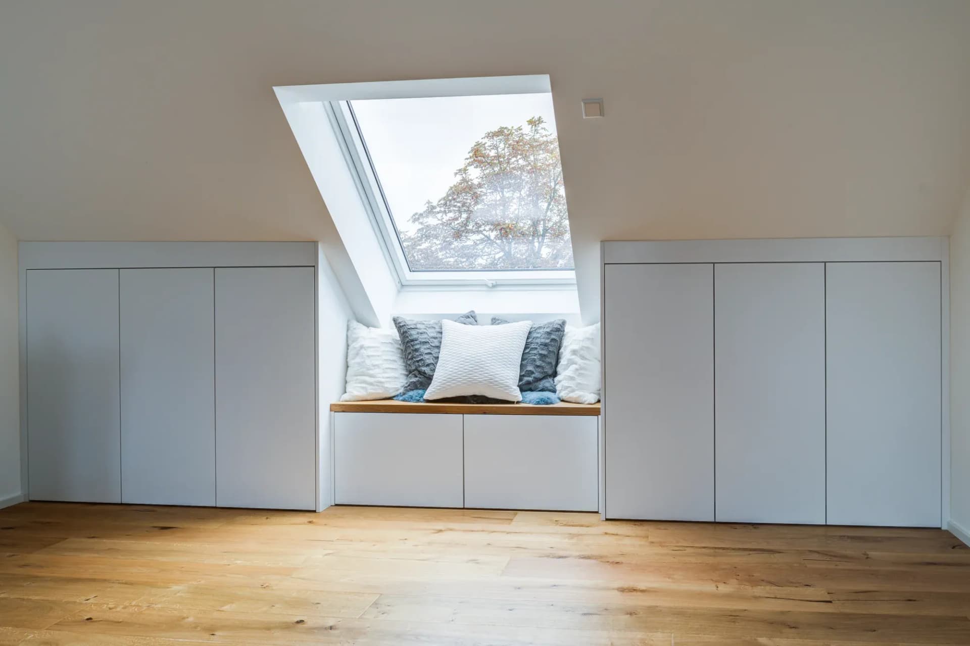 Coin banquette en chêne sous une fenêtre de toit, flanqué de placards intégrés blancs, avec un arbre visible dans la fenêtre.