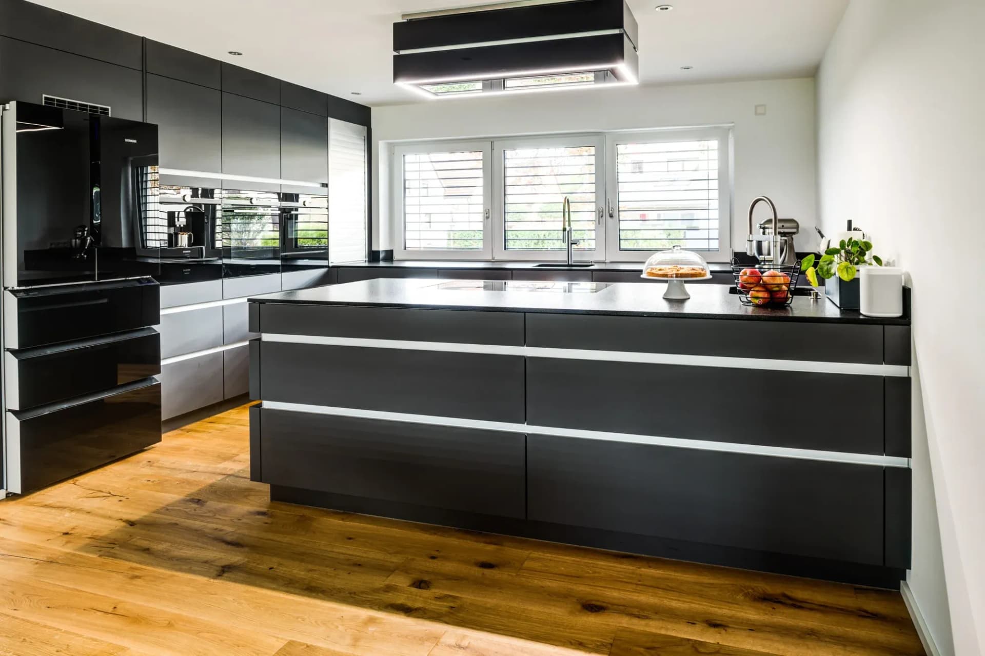 Cuisine sans poignées en anthracite et noir : îlot de cuisson autonome avec plan en granit noir, colonnes toute hauteur en arrière-plan et bandeau de fenêtres, au sol des lames de chêne.