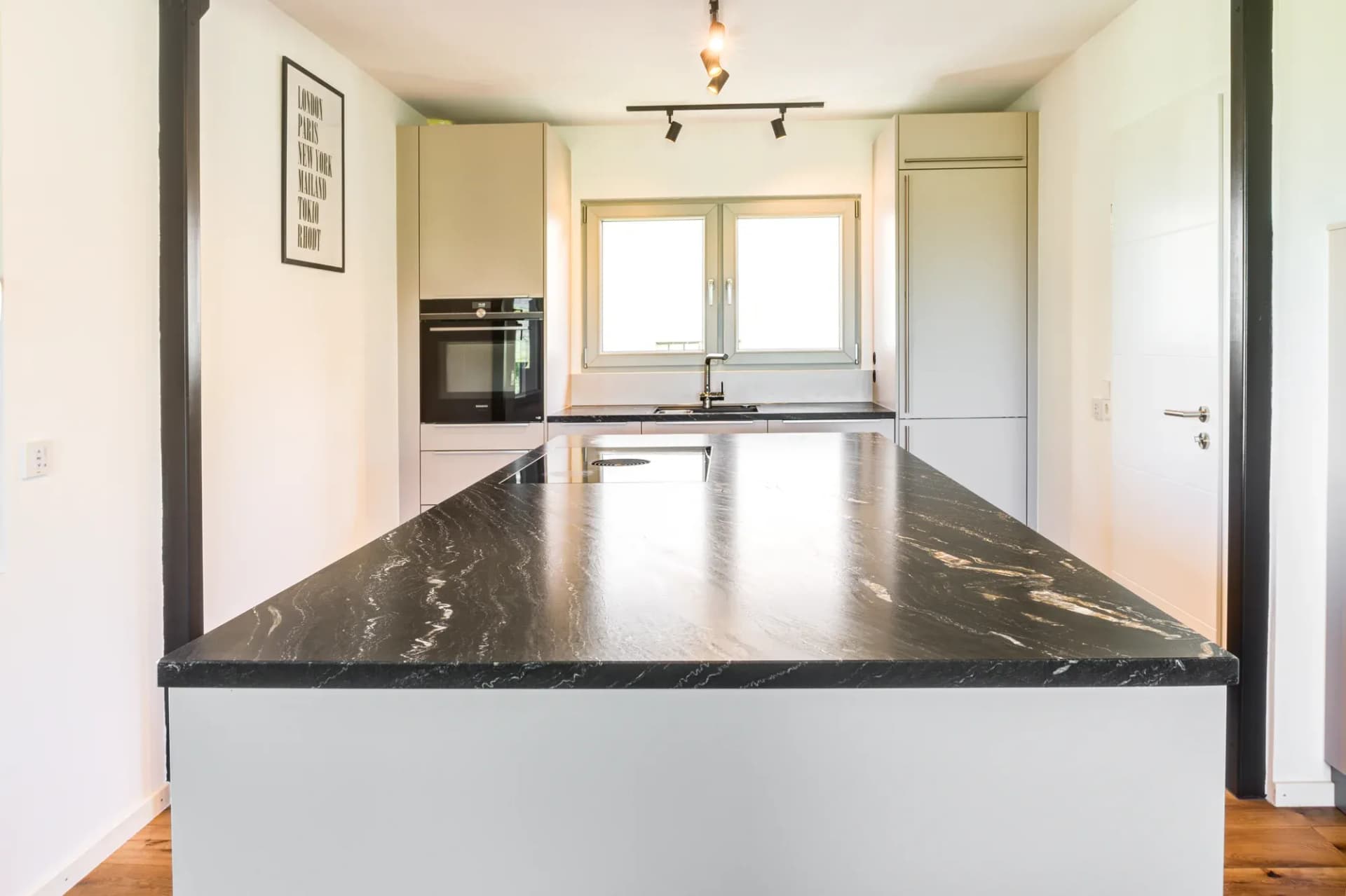 Îlot de cuisine avec plan en pierre naturelle foncée veinée de blanc, derrière la rangée de cuisine sans poignées avec four et fenêtre