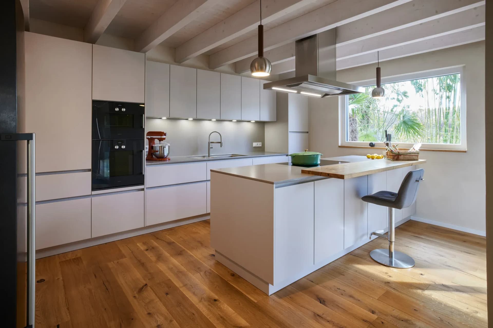 Cuisine blanche ouverte sans poignées sous plafond à poutres apparentes, avec îlot de cuisson, tabourets de bar et parquet en chêne.