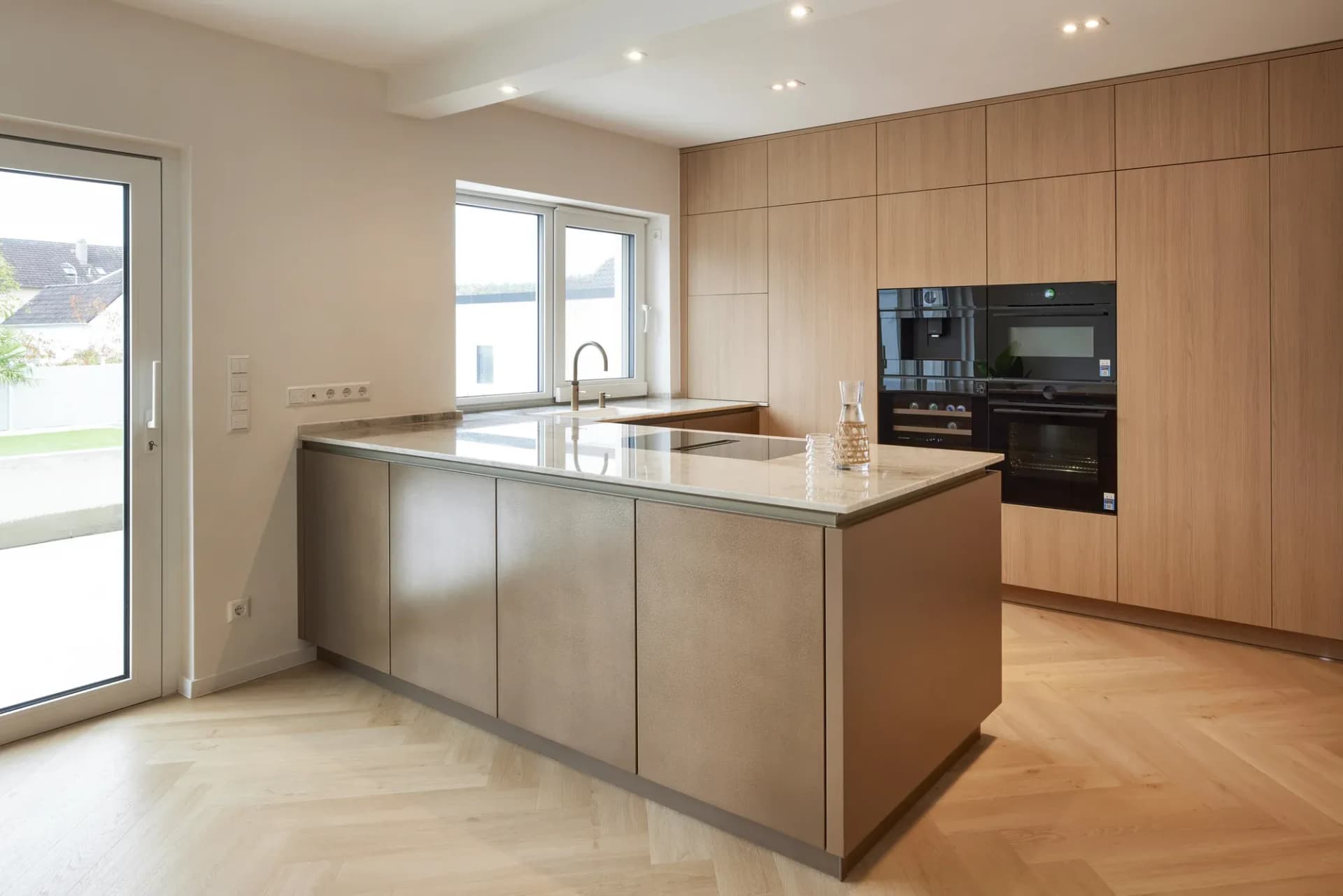 Cuisine avec îlot central ton bronze, plan de travail en pierre naturelle claire et colonnes en chêne avec électroménager encastré noir, parquet en chevrons au sol.