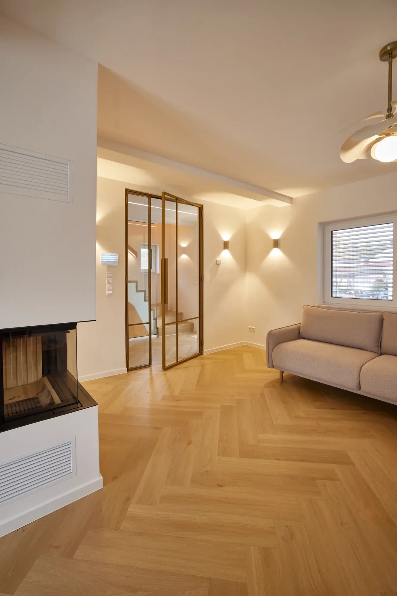Espace de vie avec cheminée blanche, porte vitrée à cadre couleur laiton menant à la cage d'escalier et canapé rembourré clair sur parquet à chevrons.