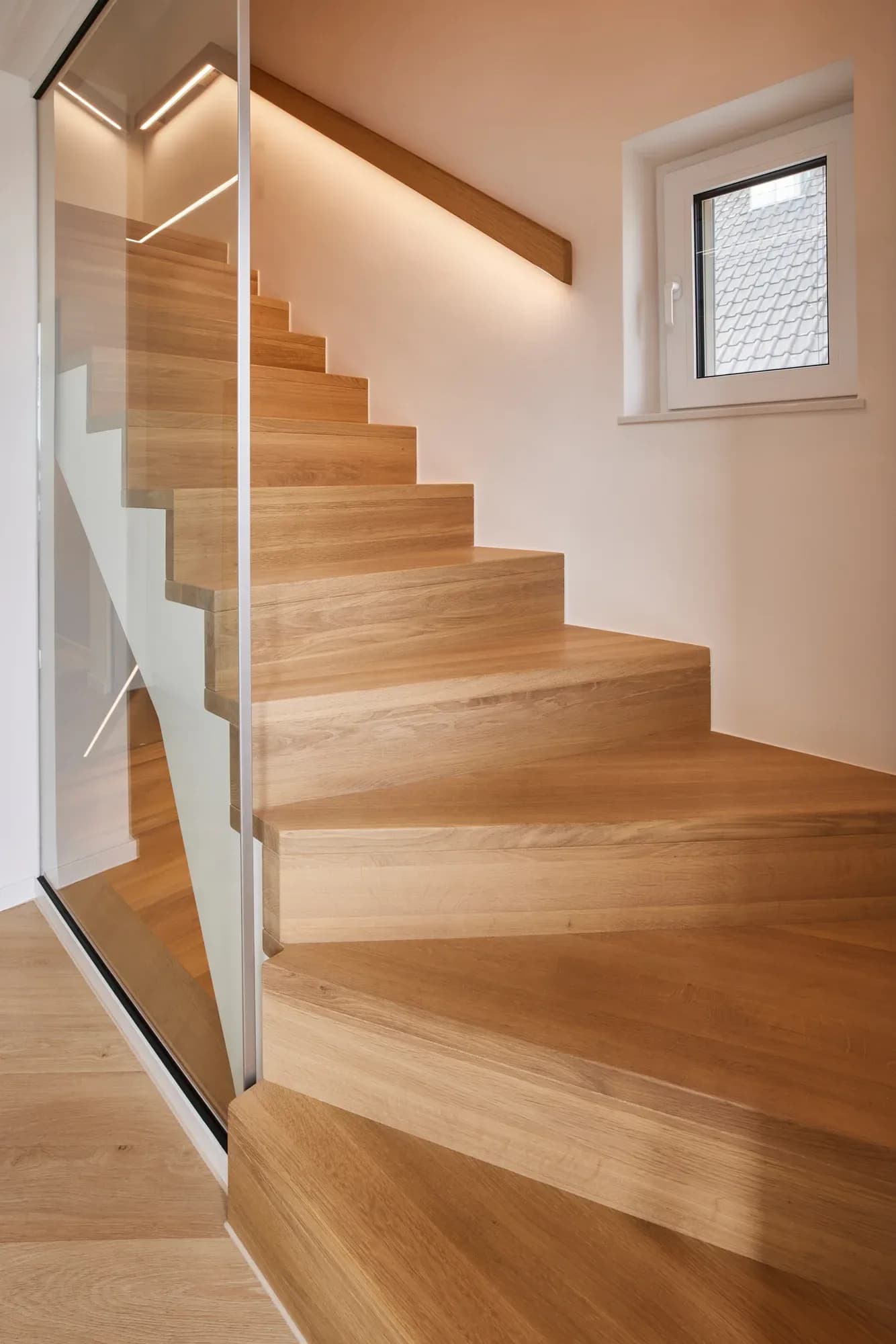 Escalier avec marches massives en chêne, paroi en verre en garde-corps et main courante murale en chêne avec éclairage LED.