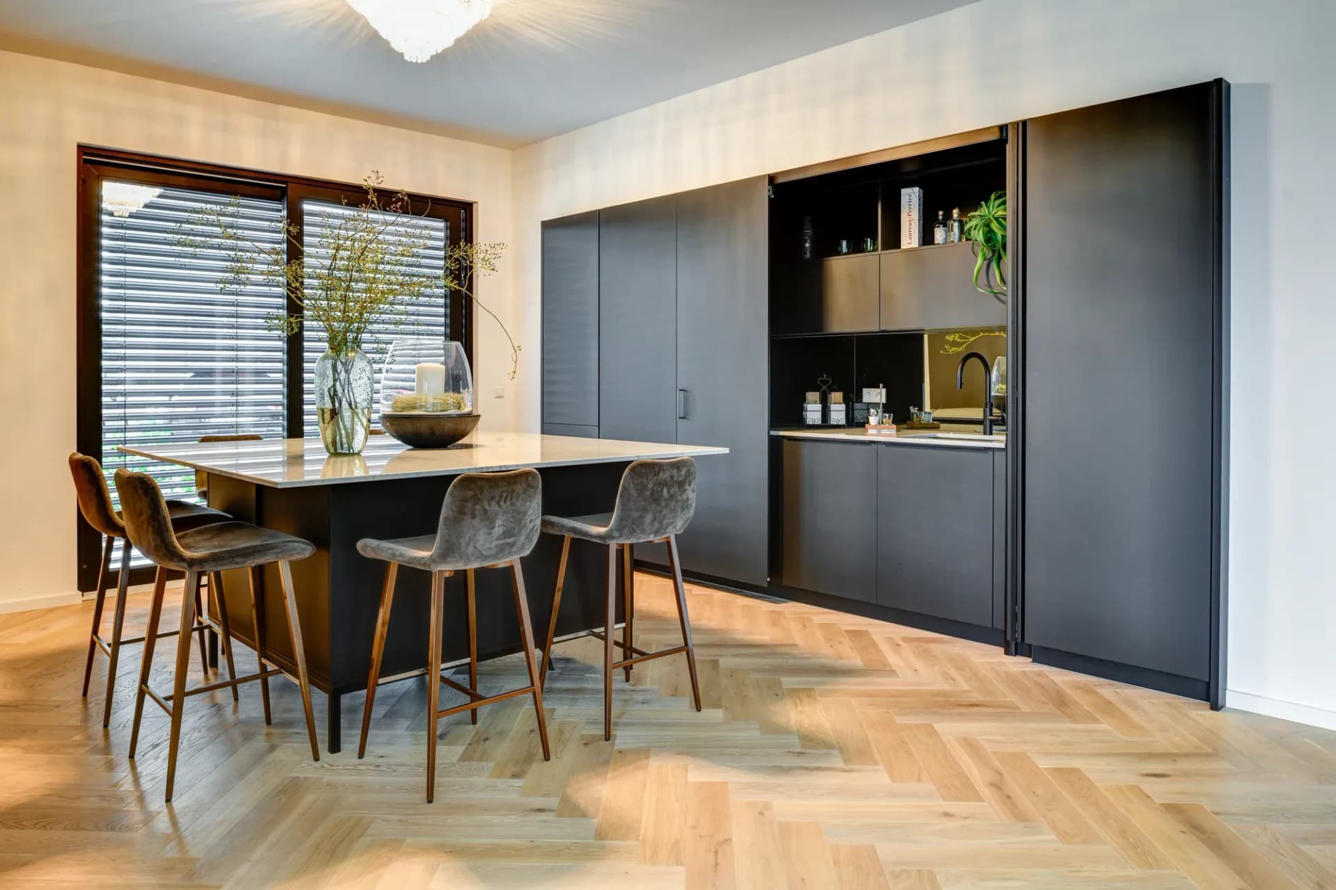 Cuisine de réunion avec façades anthracite sans poignées, îlot en pierre naturelle claire, tabourets de bar en cuir et stores en bois au-dessus d'un sol en chêne posé à bâtons rompus.