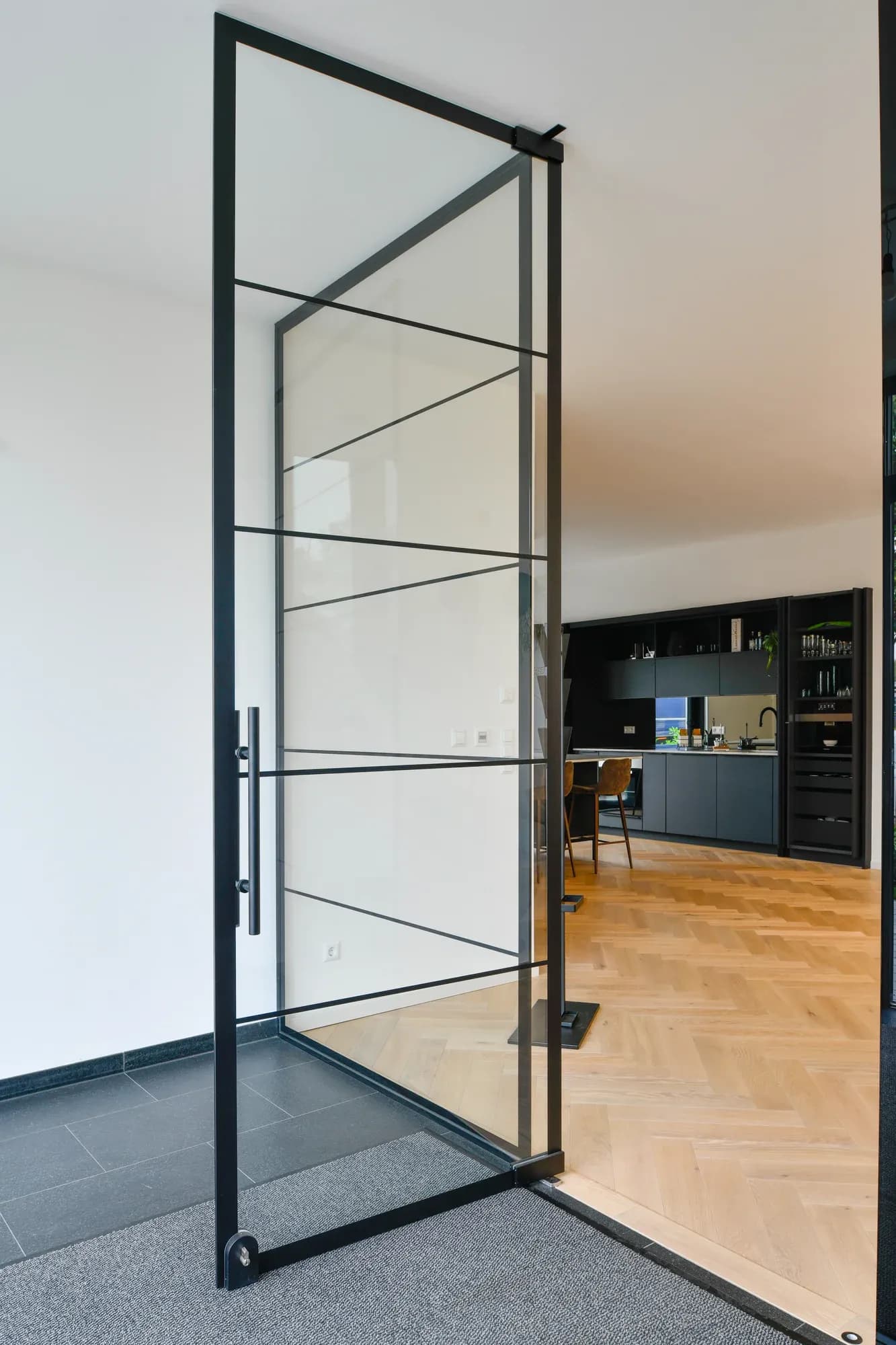 Porte loft en cadre acier noir et verre, menant de l'entrée au sol en ardoise vers les bureaux au parquet en chevrons.