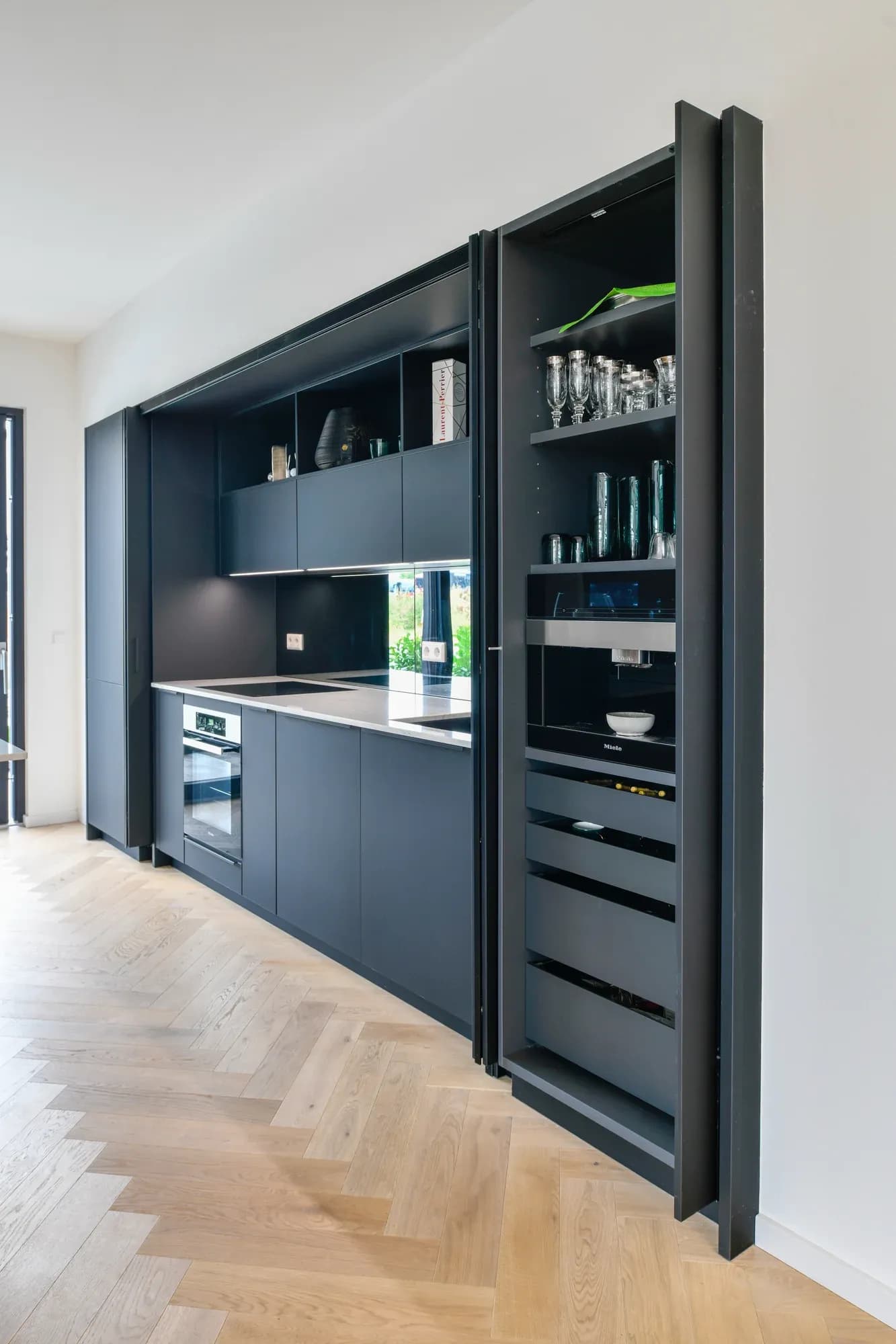 Cuisine linéaire entièrement intégrée en anthracite avec armoire de rangement à porte escamotable ouverte : verres, machine à café automatique Miele et tiroirs.
