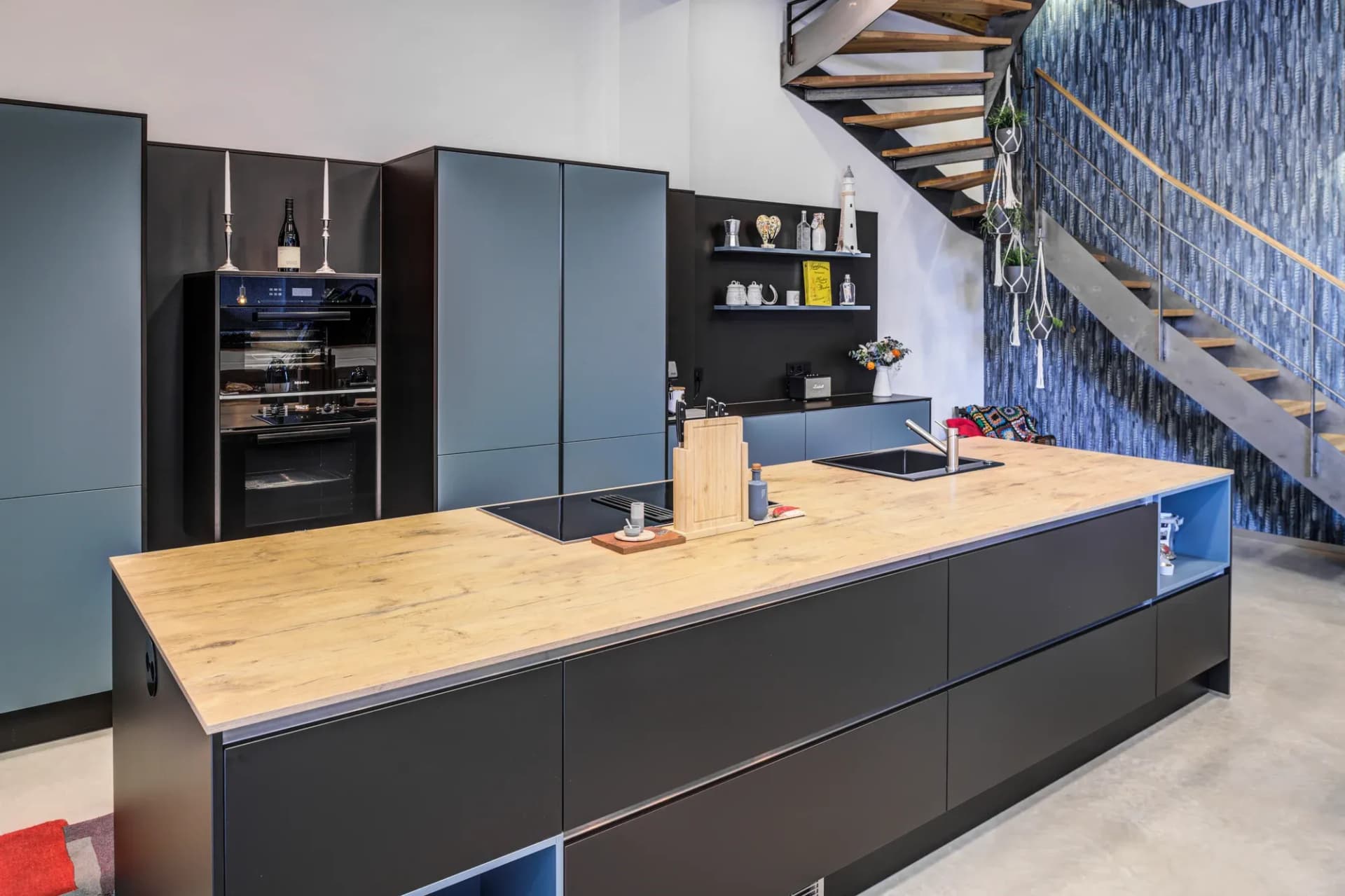 Cuisine ouverte bleu pétrole et noir mat avec plan de travail en chêne massif, avec derrière un escalier à limon en acier plat devant un mur à motifs bleus.