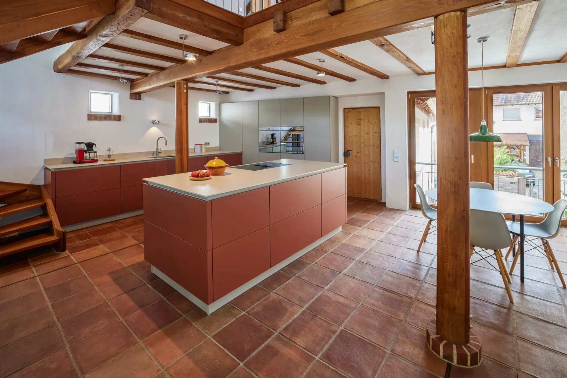 Cuisine ouverte dans une maison à colombages : îlot indépendant aux façades sans poignées rouge terre cuite, accordées au sol en carreaux de terre cuite ; derrière, des colonnes vert sauge et un coin repas sous les poutres apparentes.
