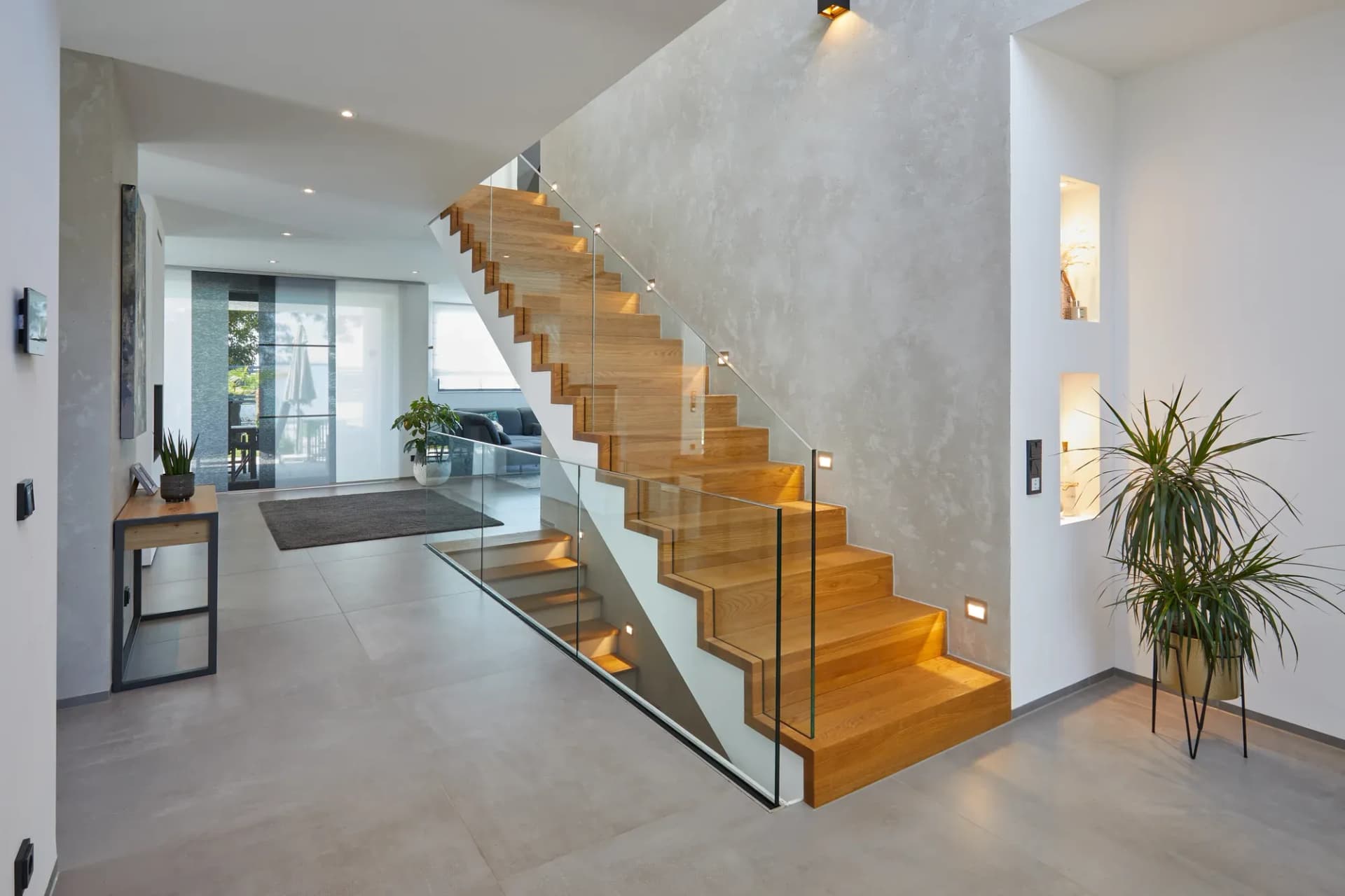 Escalier plié en chêne, sans structure visible, avec garde-corps en verre devant un mur effet béton, dans l'entrée ouverte avec vue sur le séjour.