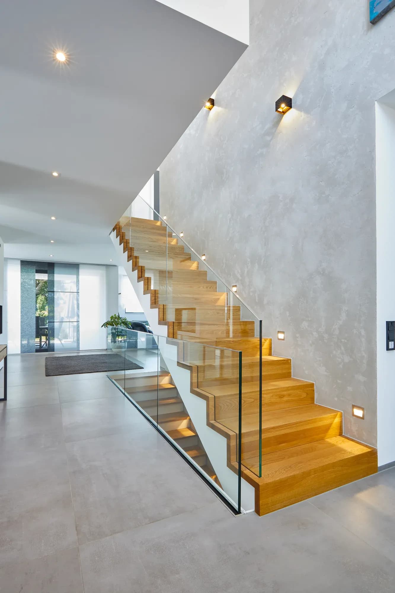 L'escalier en chêne vu d'en bas : marches pliées, garde-corps en verre et éclairage de marches encastré dans le mur.
