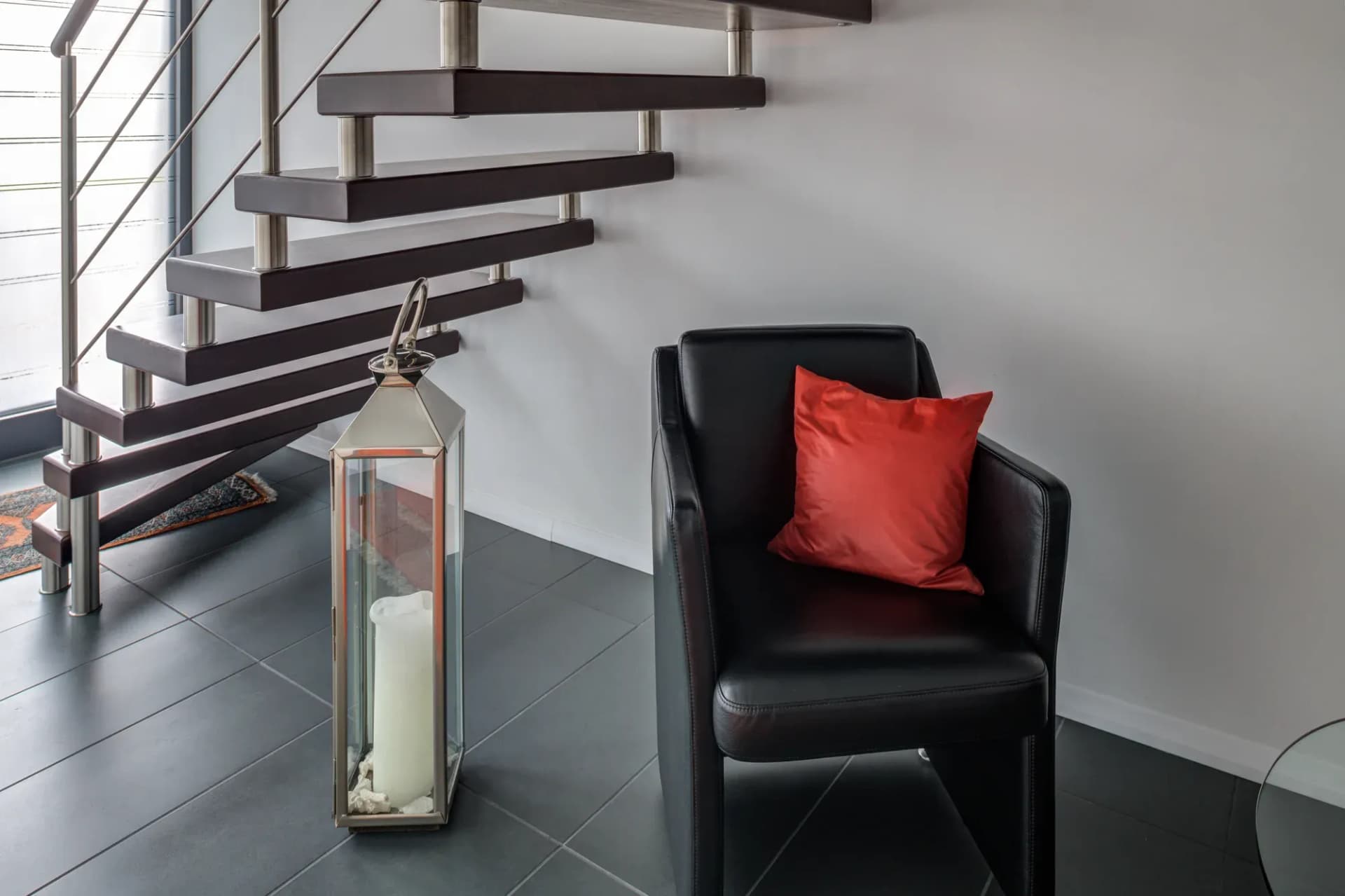 Escalier à boulons aux marches couleur acajou et garde-corps en acier inoxydable, devant un fauteuil en cuir et un coussin rouge.