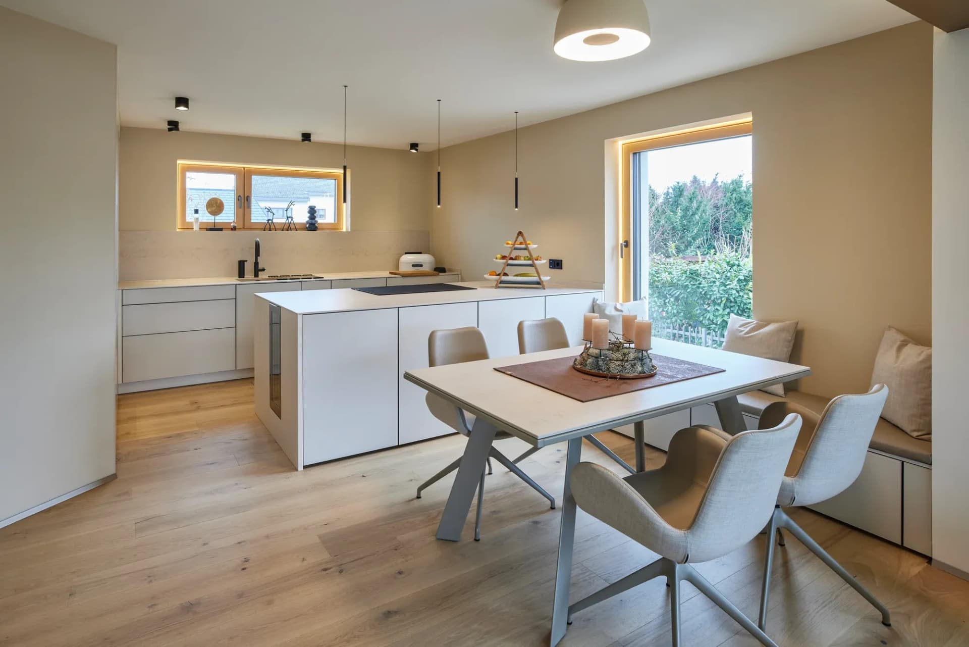 Espace de vie ouvert cuisine-séjour-repas avec cuisine blanche sans poignées, îlot de cuisson et parquet en chêne clair ; à droite, la table à manger avec chaises rembourrées et une banquette dans la niche de la fenêtre.