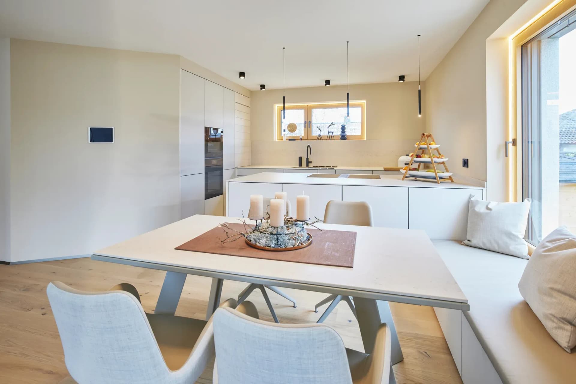 Vue sur la table à manger dressée avec bougies vers la ligne de cuisine blanche ; murs chauds couleur sable, sol en chêne et fenêtre toute hauteur avec banquette.