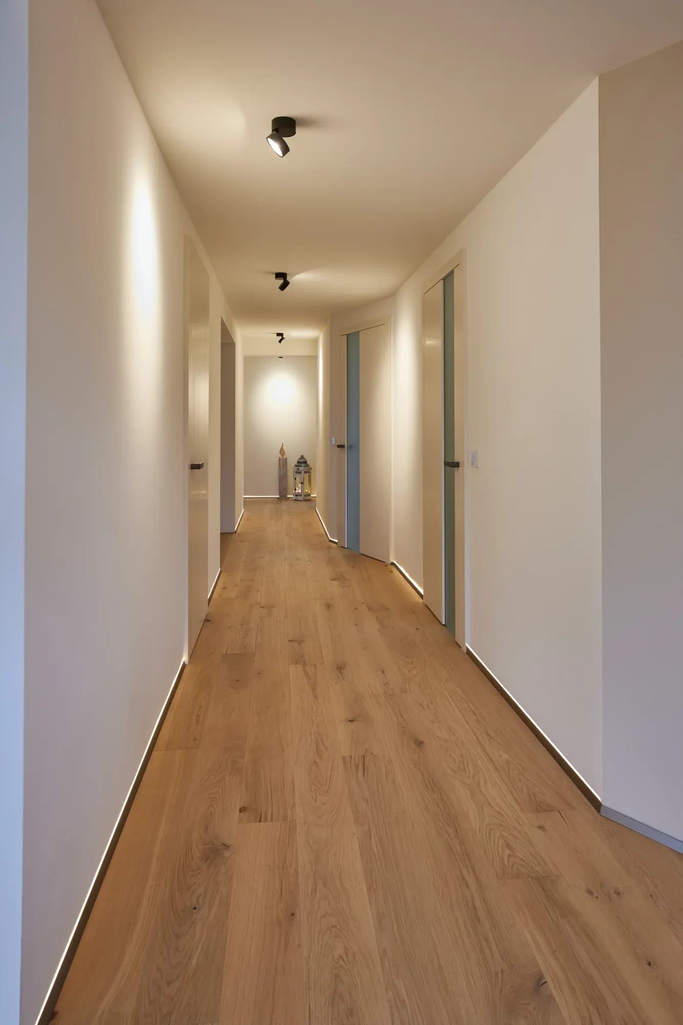 Couloir avec portes intérieures claires et parquet en chêne continu ; éclairage indirect sur les murs et fente lumineuse au ras du sol.