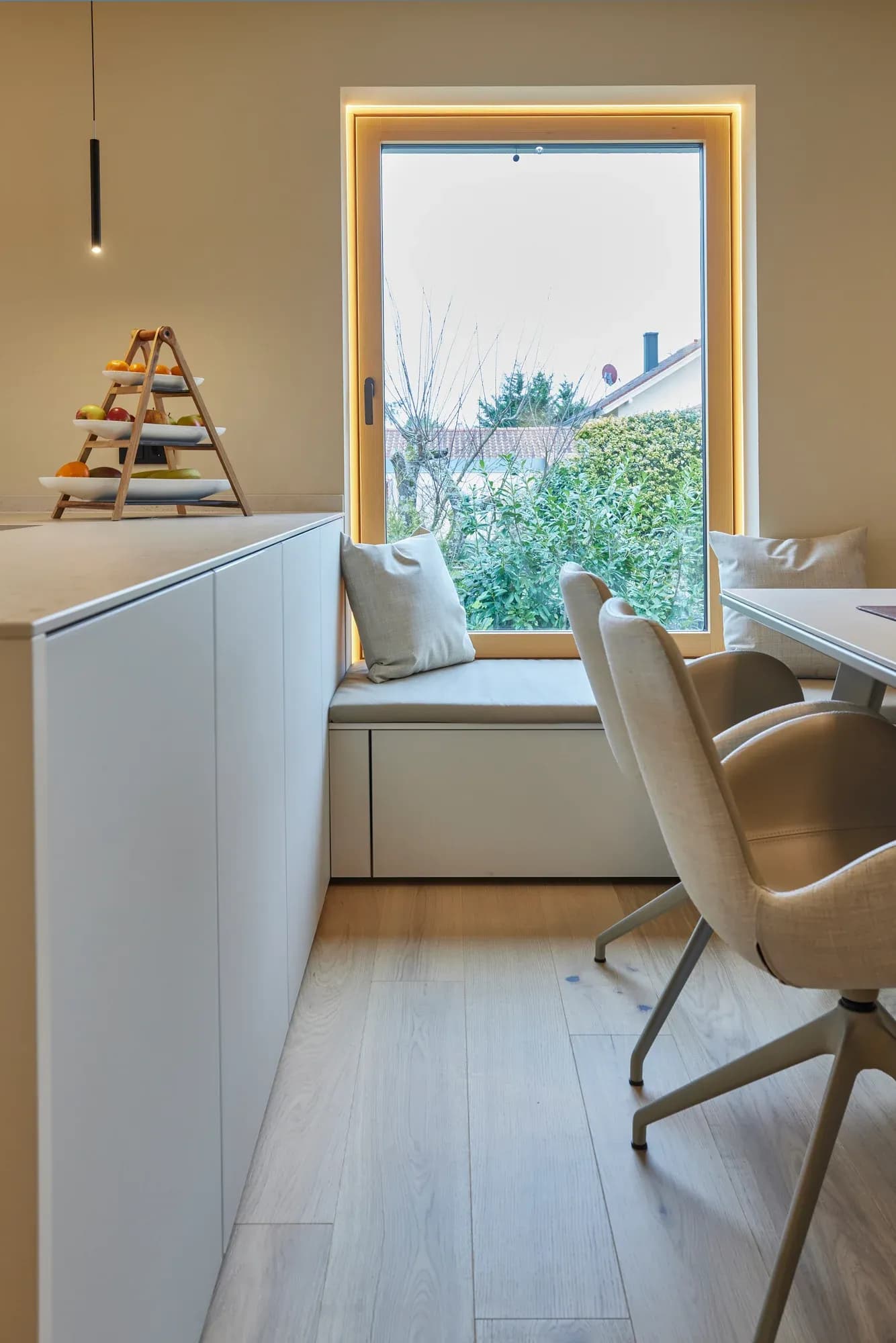 Banquette rembourrée dans la niche de fenêtre avec embrasure à éclairage indirect ; devant, une chaise rembourrée claire à la table à manger, à côté une rangée de buffets blancs.