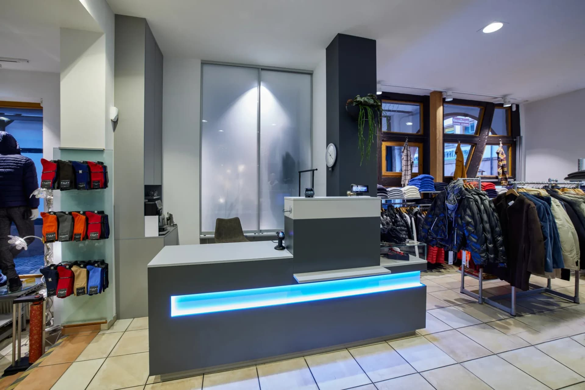 Comptoir de vente anthracite avec plan de travail clair et ligne lumineuse LED bleue dans un magasin de mode masculine, portants à vêtements en arrière-plan.