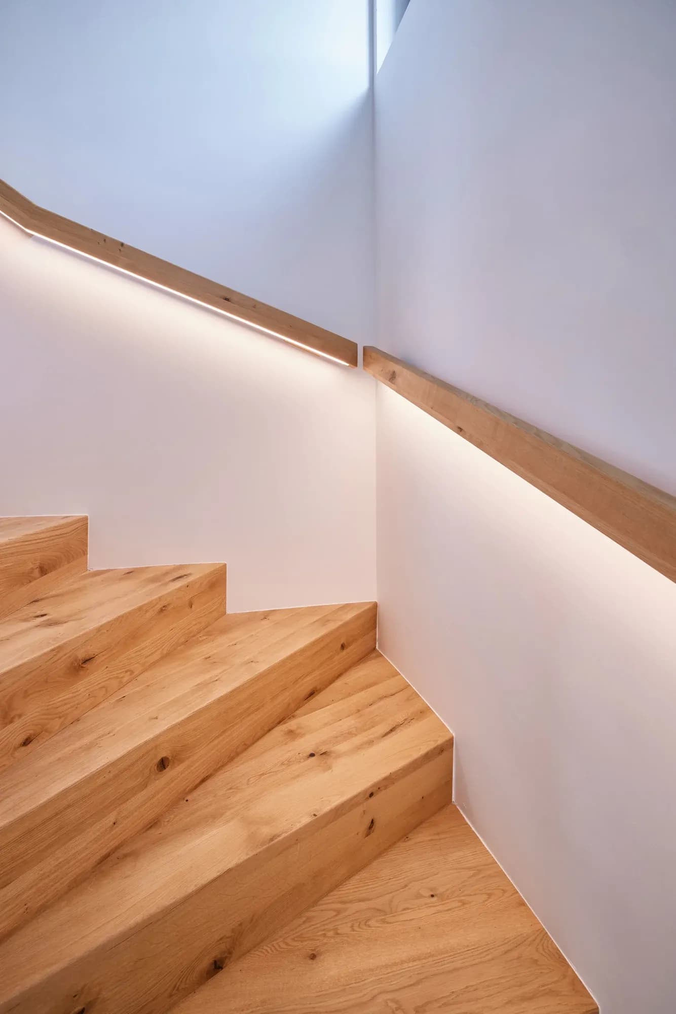 Escalier en bois massif avec main courante rétroéclairée, qui guide la lumière le long de l'angle du mur.